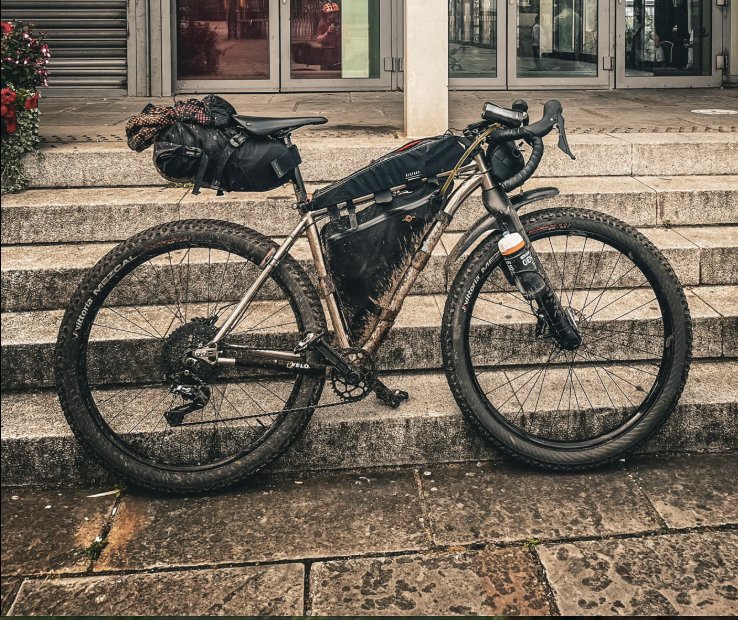 A standing gravel bike loaded with bikepacking gear, including a rear saddlebag and frame bags, positioned on stone steps with a building entrance in the background.