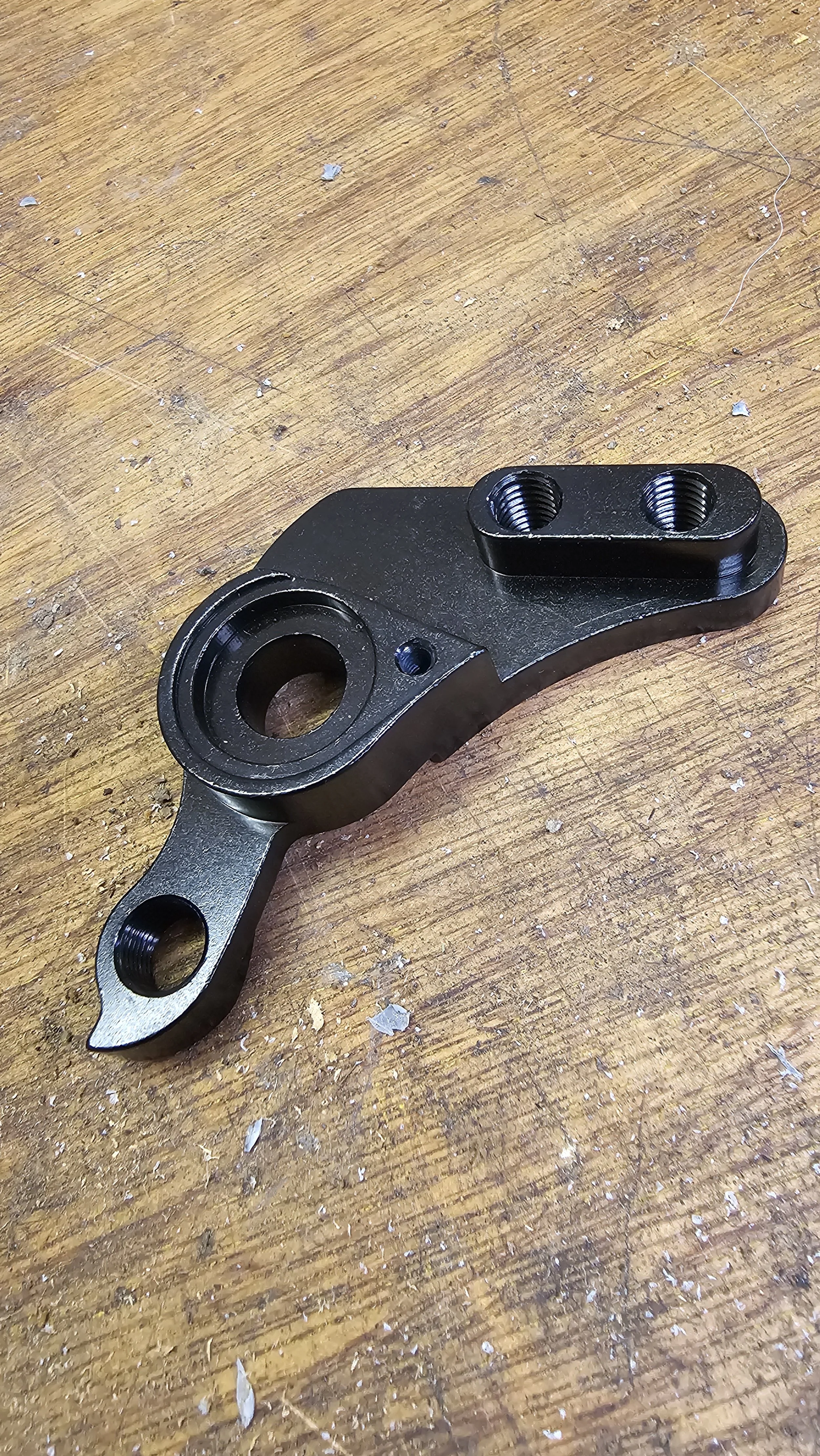 Black angular metal component with multiple threaded holes, placed on a wooden surface.