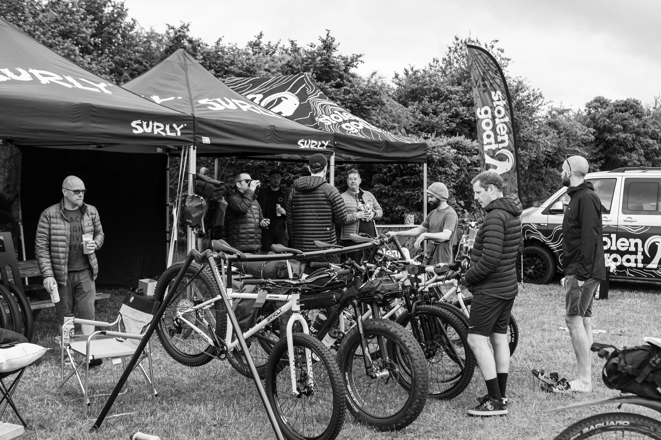 Group of people gathered at an outdoor event with bicycles, tents, and a van, some talking and some holding drinks, with a wooded background.