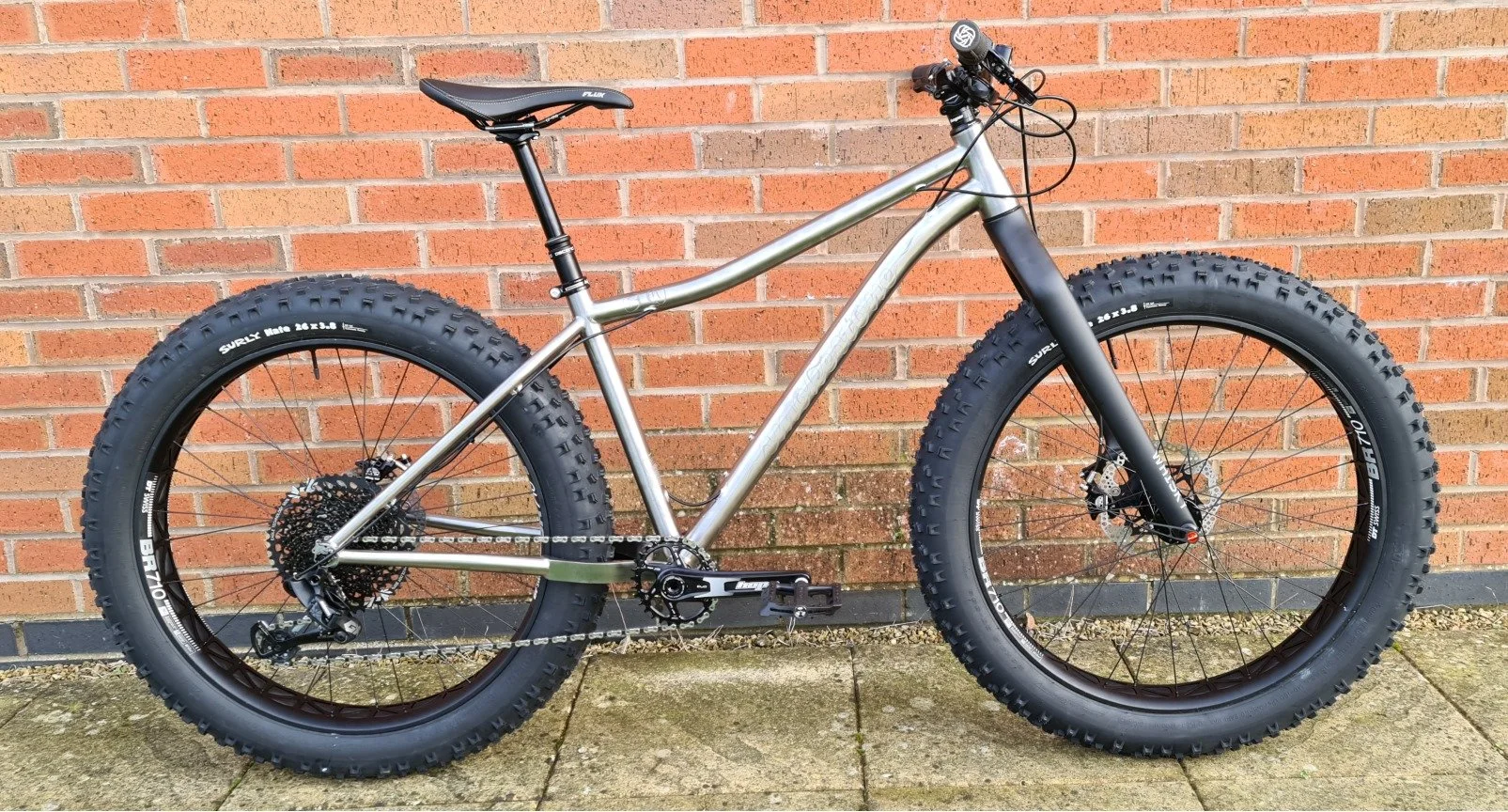 A silver fat-tire mountain bike with black front suspension fork, black saddle, and knobby tires, leaning against a red brick wall outdoors.