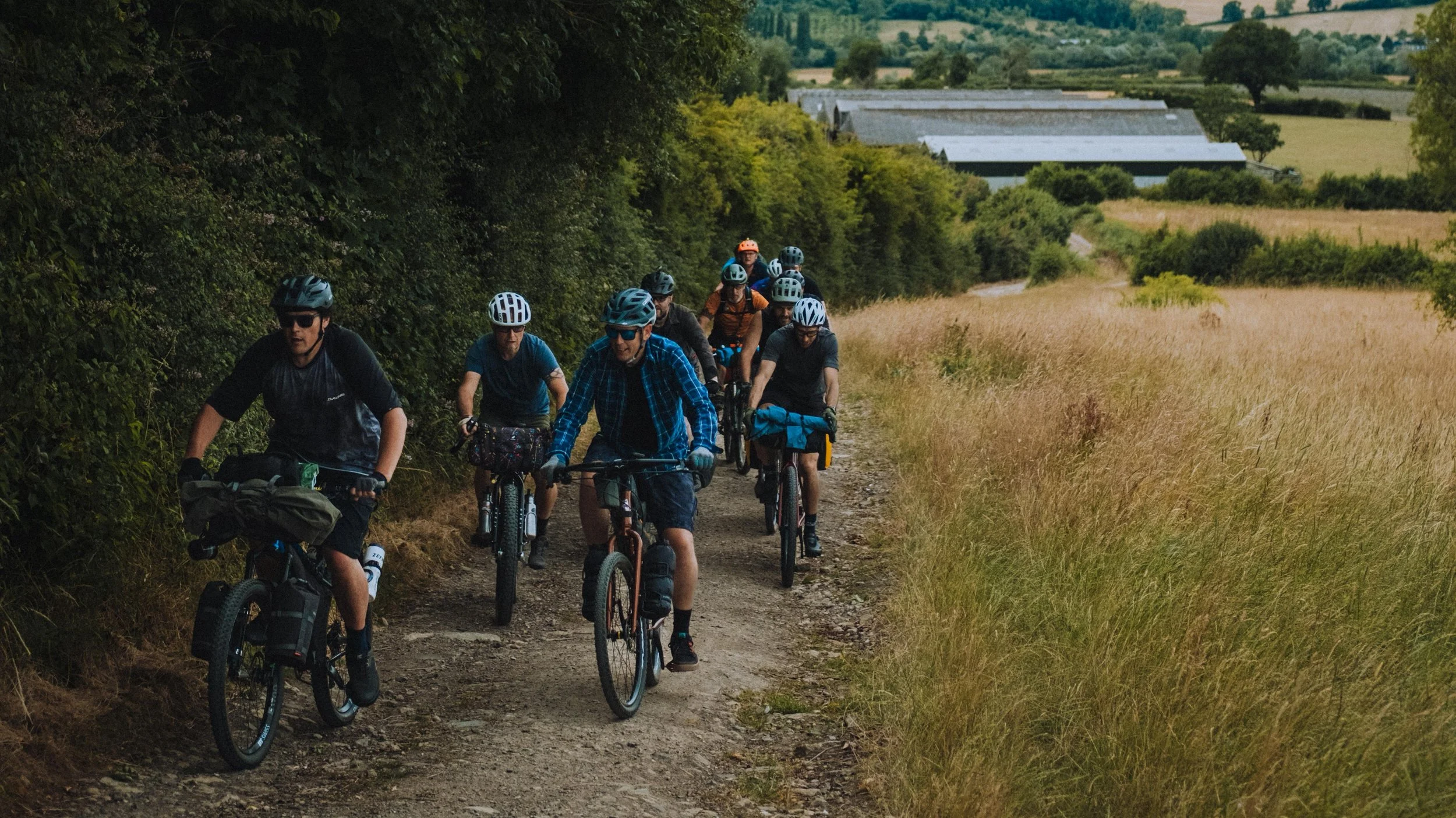 A group of people biking along a dirt trail through a rural area, with fields and trees on either side and farm buildings in the background.