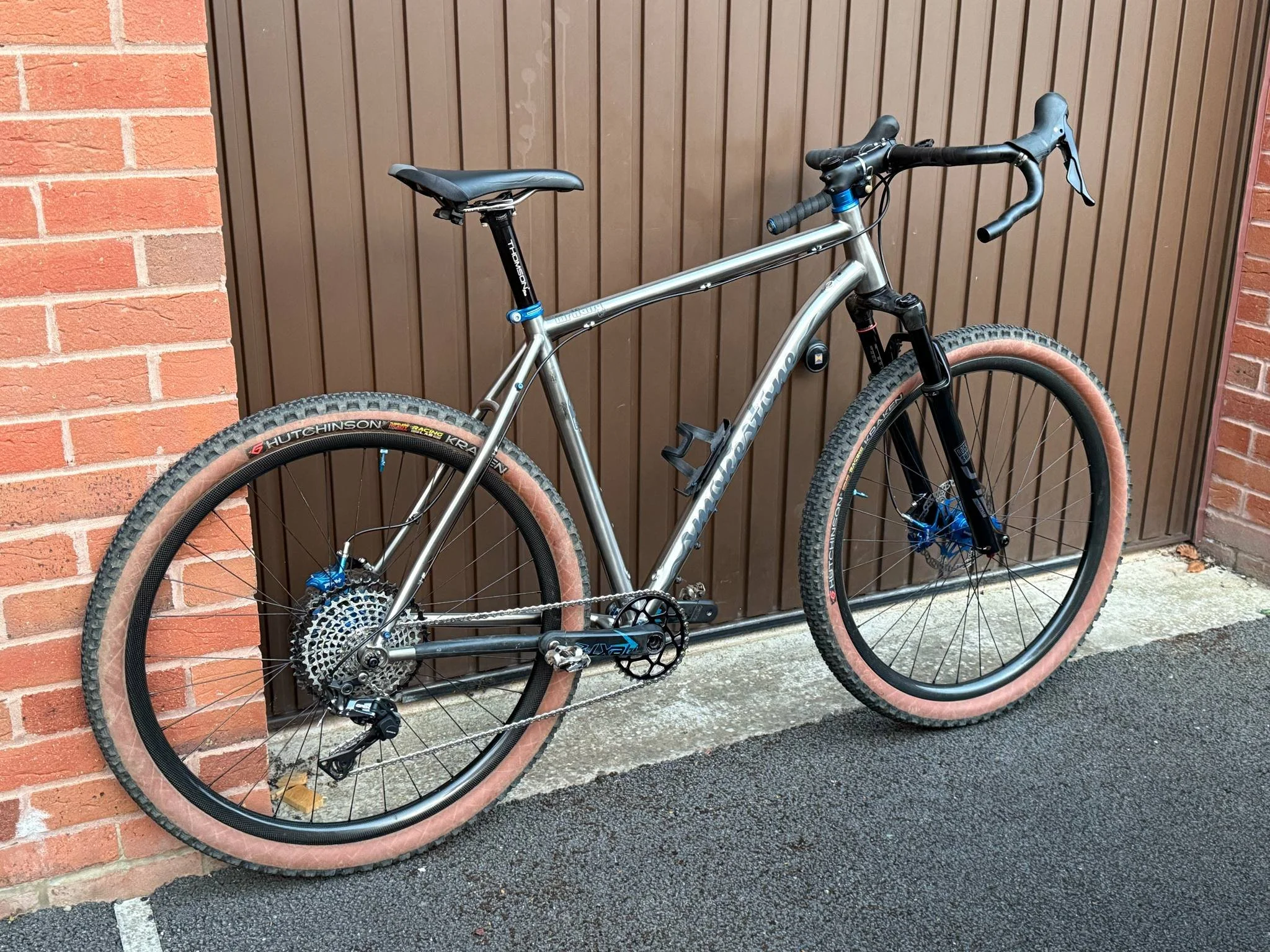 A mountain bike with a metallic frame and knobby tires is leaning against a brown wooden door on a brick and concrete surface.