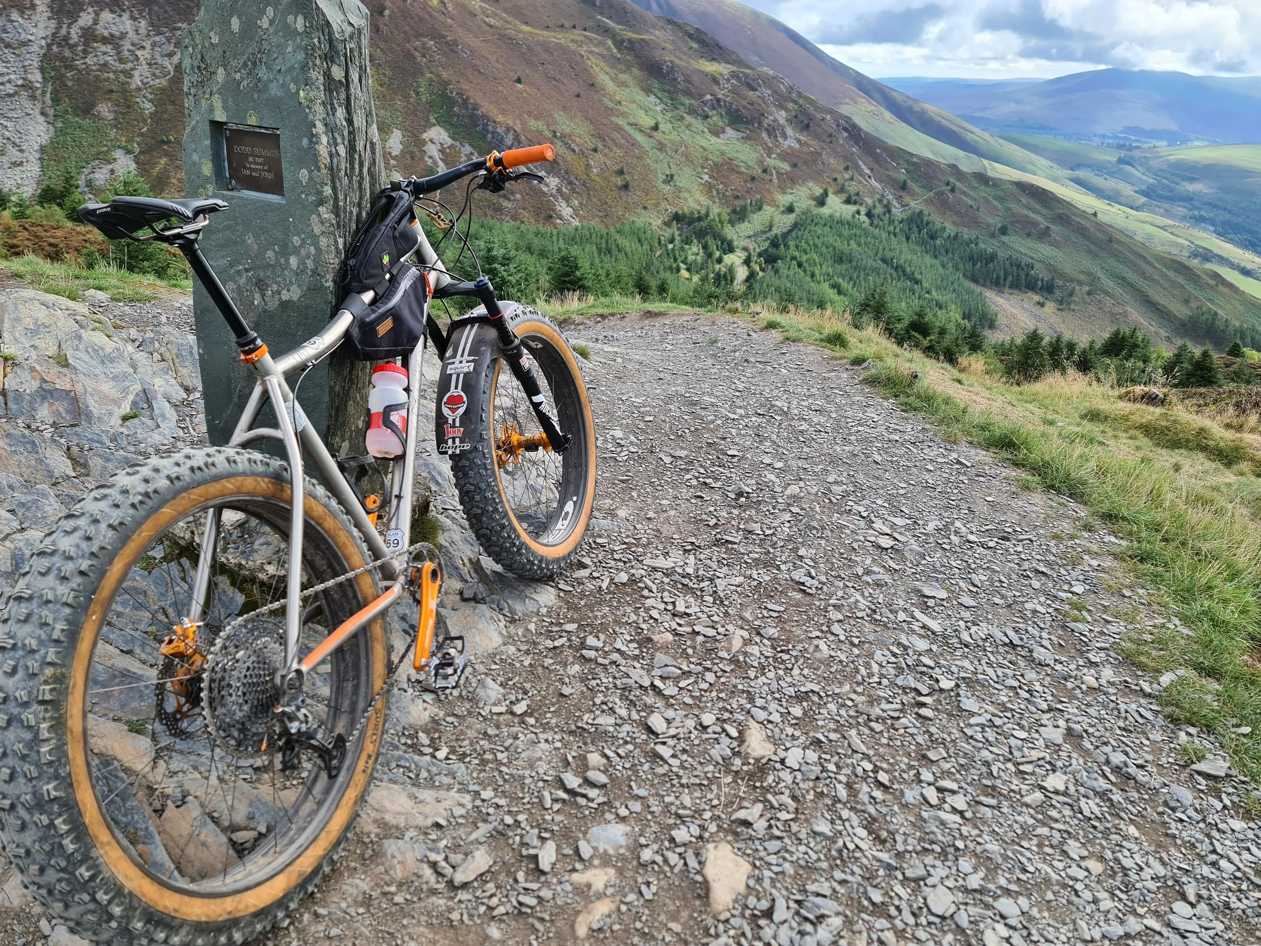 Fat bike leaning against a stone marker on a gravel mountain trail with a scenic view of hills and valleys in the background.