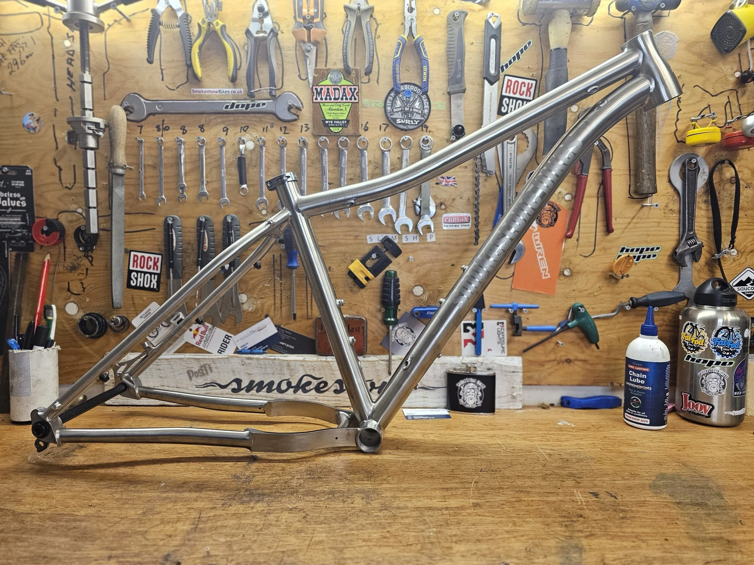 Steel bicycle frame on a workbench surrounded by various tools and equipment, including wrenches, pliers, and lubricant in a workshop setting.
