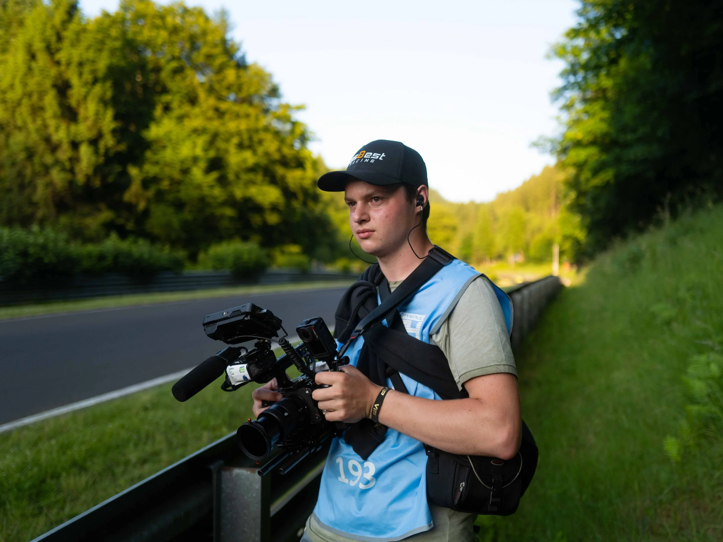 A young man holding a professional video camera on the side of a racetrack surrounded by trees.