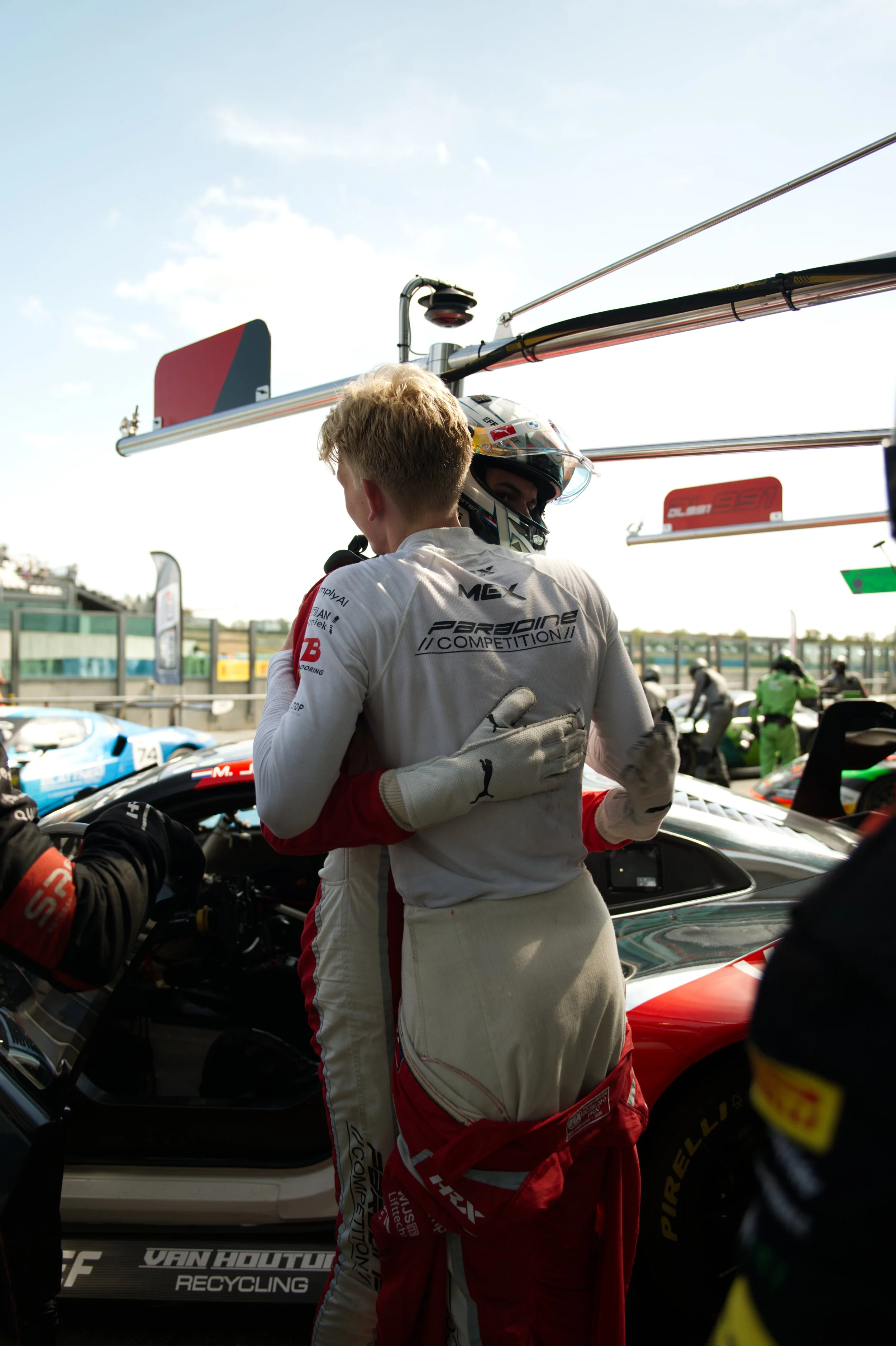 Two race car drivers are hugging in a garage area surrounded by racing cars and team members, with equipment and pit lane visible in the background.