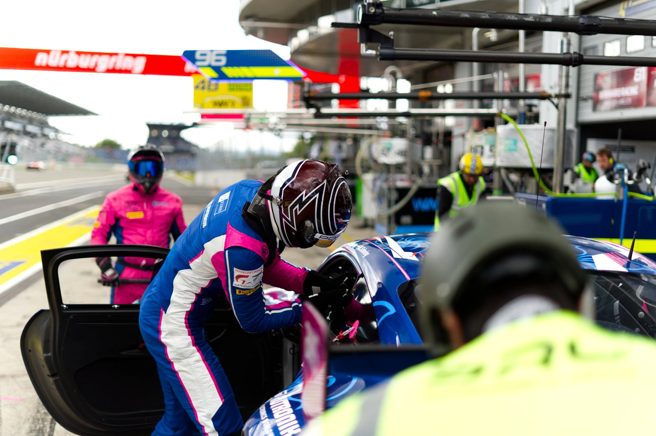 Race car driver in blue and pink racing suit enters the cockpit of a race car in the pit lane during a race event, with team members and crew working around the vehicle.