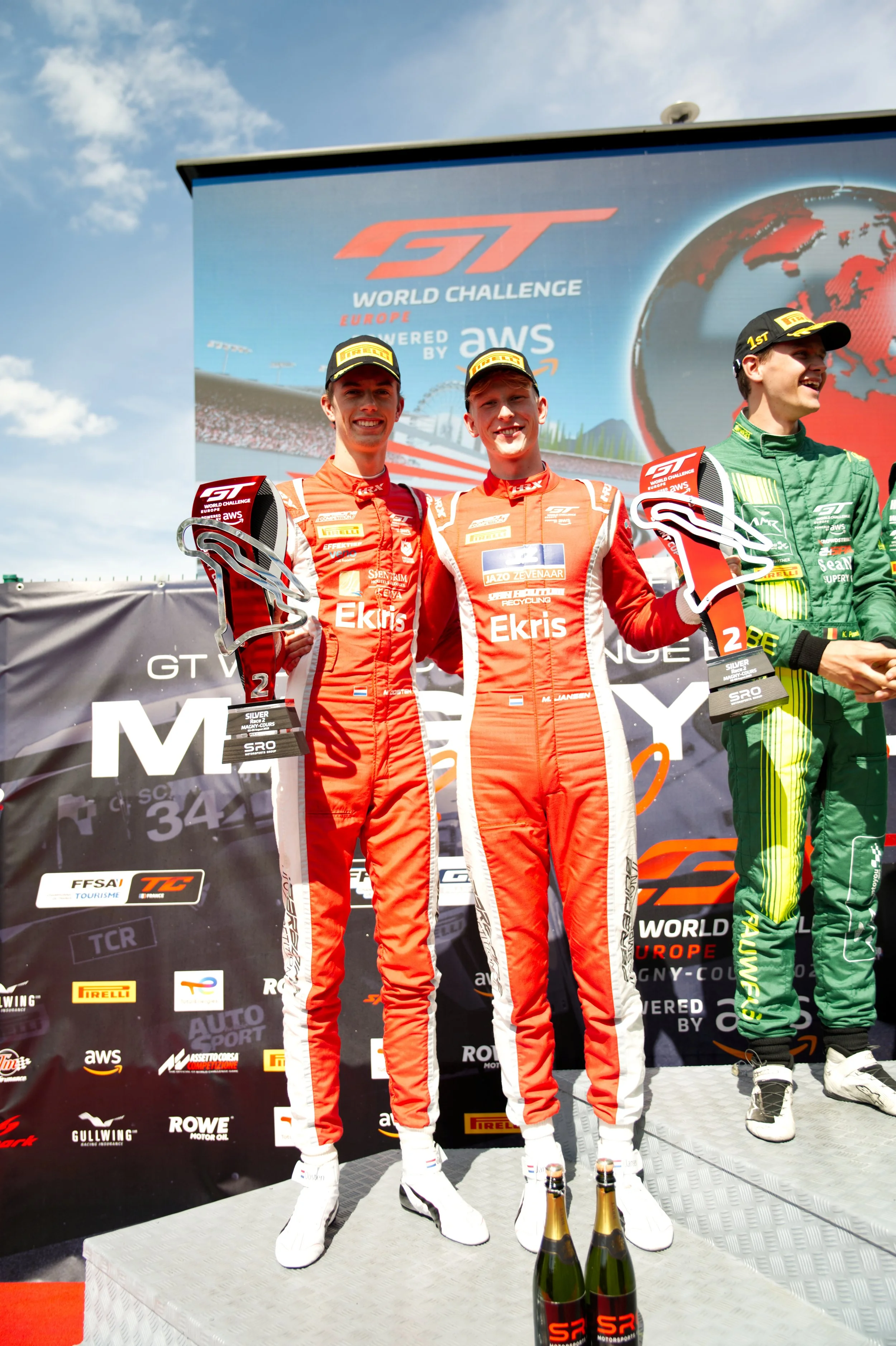 Three race car drivers on a podium celebrating their victory in the GT World Challenge Europe, with the two in orange suits holding trophies, and the other in a green suit, against a backdrop with logos and a globe.
