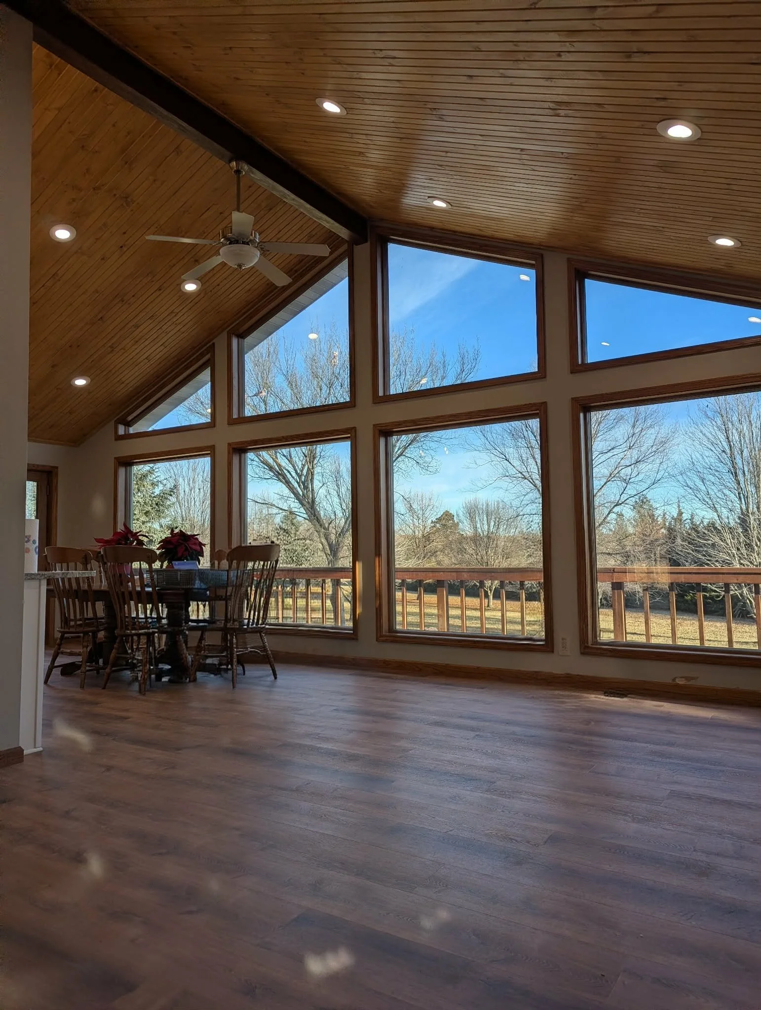 Interior view of a bright room with large windows, wooden ceiling, ceiling fan, wooden dining table with chairs, and a view of trees and blue sky outside.