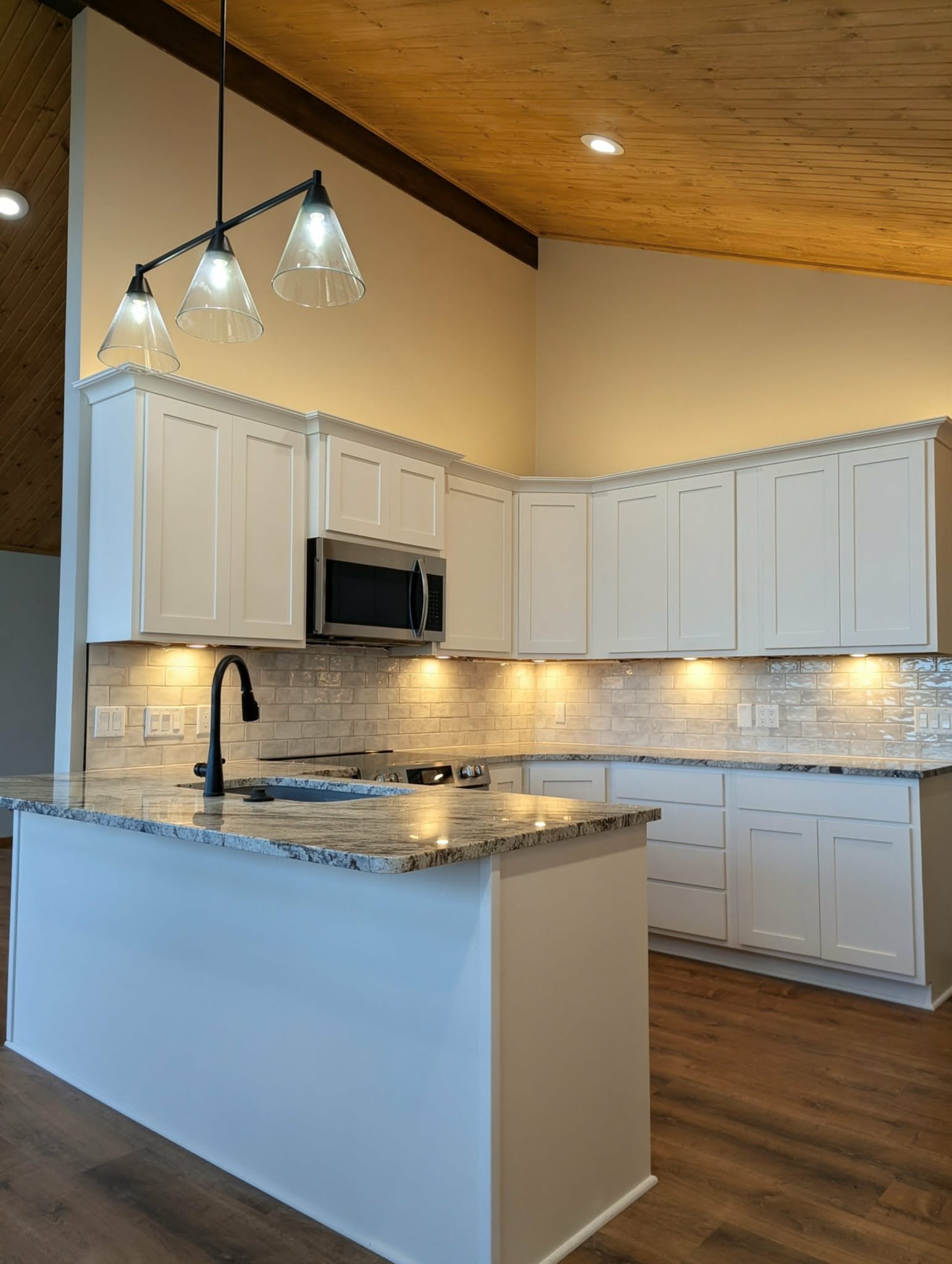 A modern kitchen with white cabinets, granite countertops, a black faucet, and a tiled backsplash, with recessed lighting and a wooden ceiling.