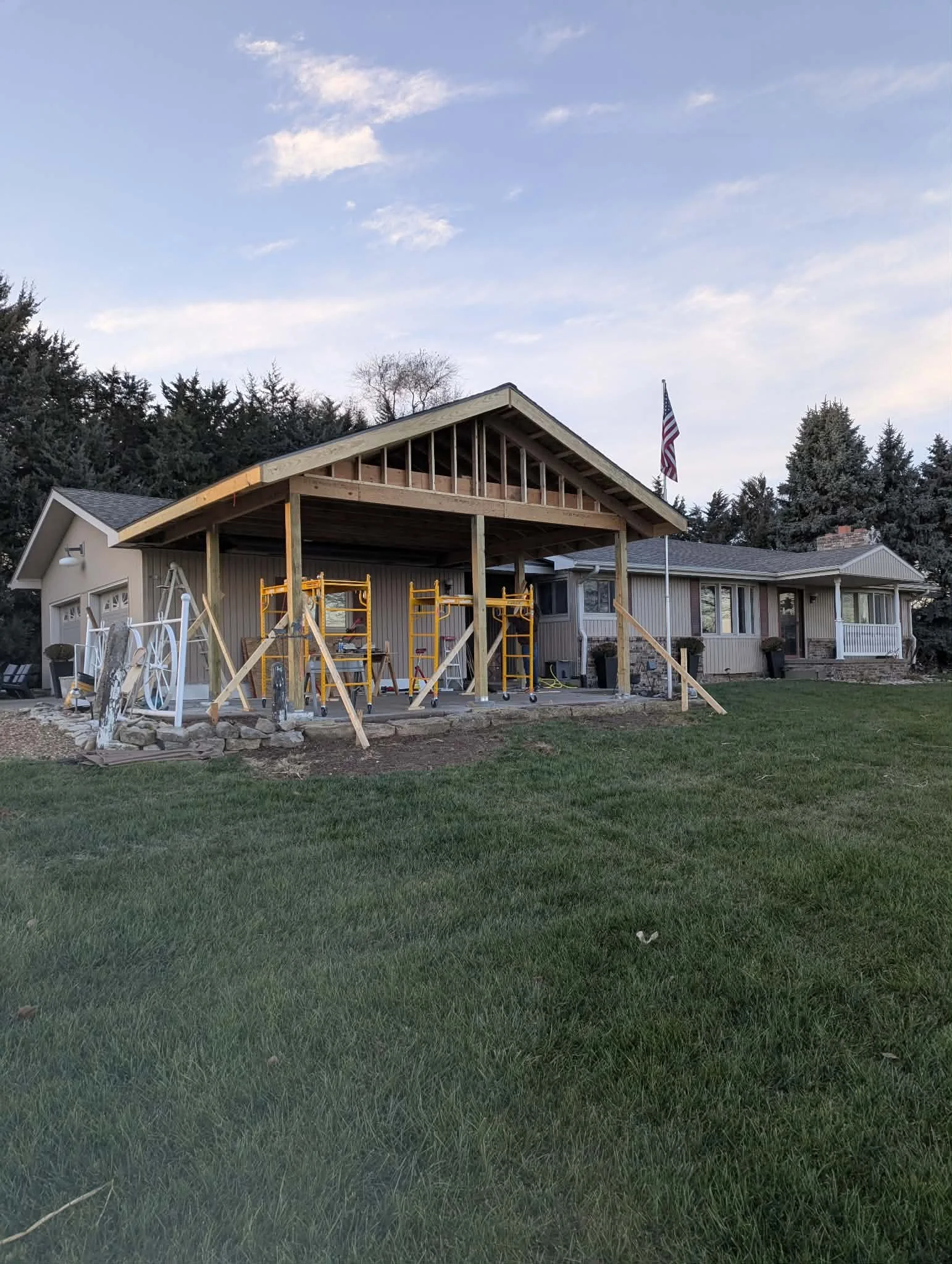 House with a patio under construction, with scaffolding and wooden framework, a lawn in the foreground, and an American flag flying in the background.