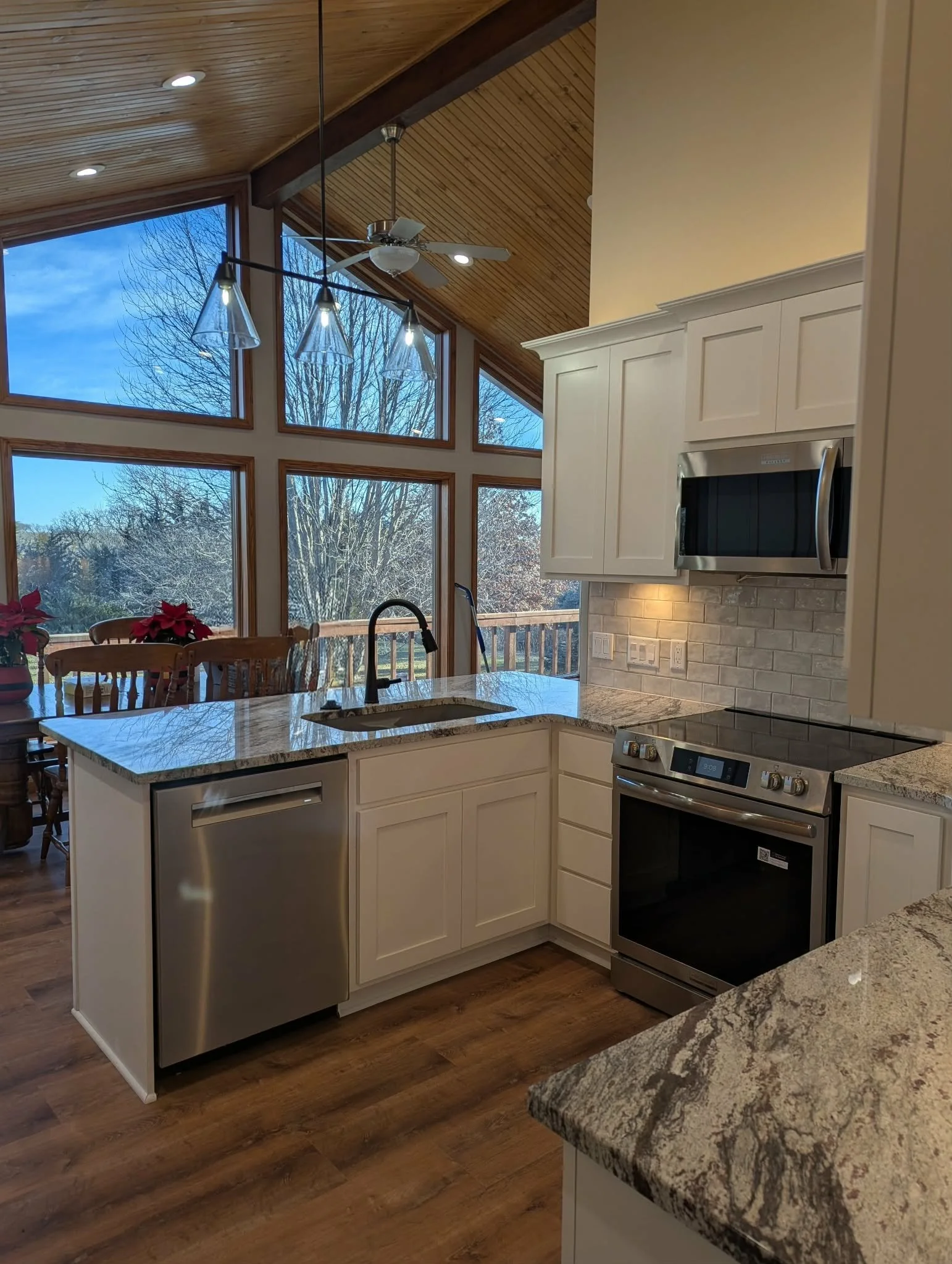 Modern kitchen with granite countertops, white cabinets, stainless steel appliances, and large windows showing a view of trees outside.