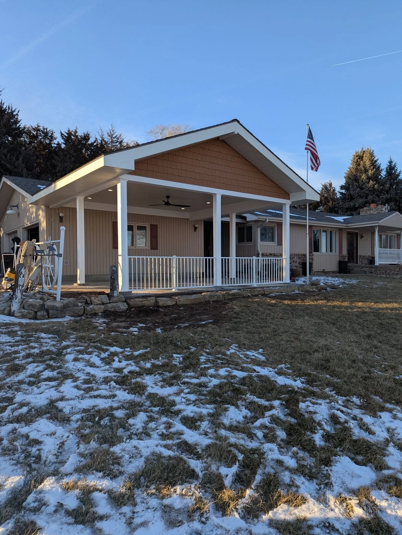 Front porch of a house with a white railing, beige siding, and a gable roof, with an American flag on a flagpole to the right, and snow on the lawn in the foreground.