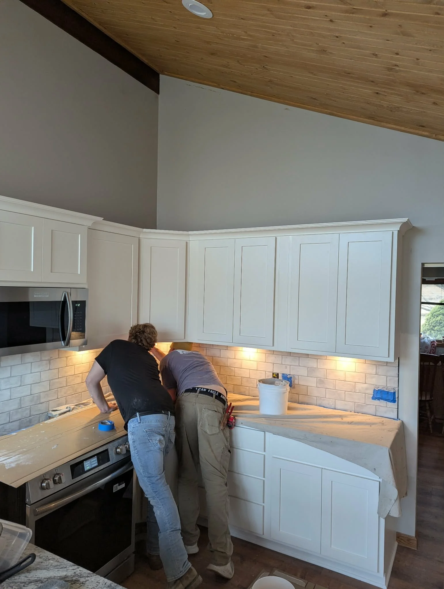 Two men working on installing white kitchen cabinets and backsplash in a home renovation, with tools and materials on the countertop.