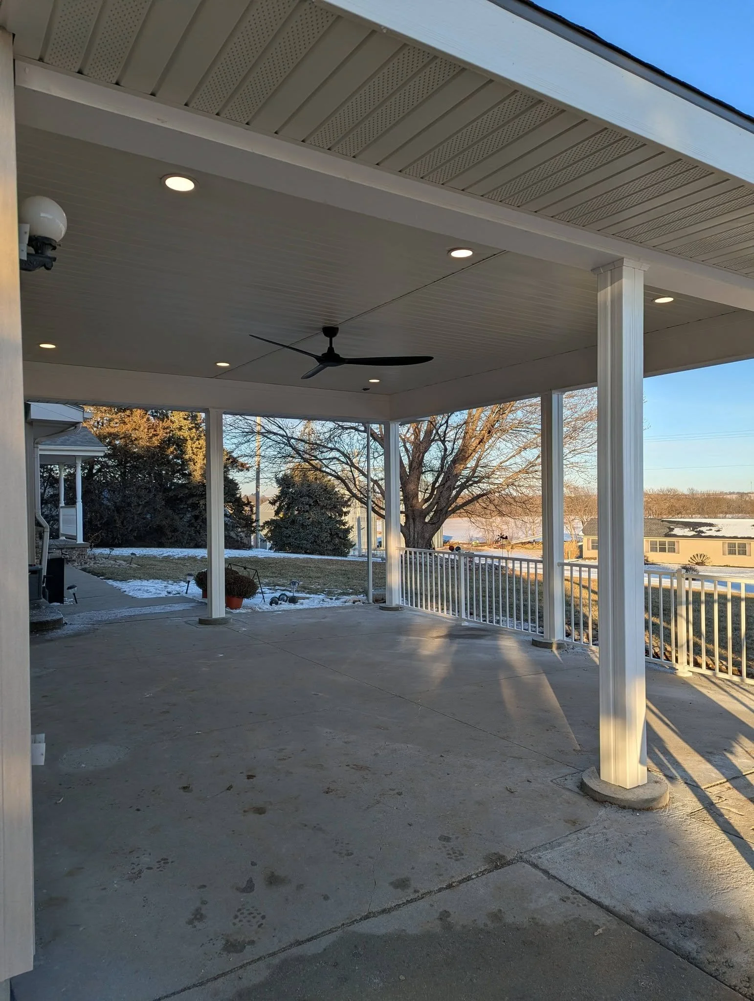 Covered porch with ceiling fan, recessed lighting, and white railing, overlooking a yard with trees and snow.