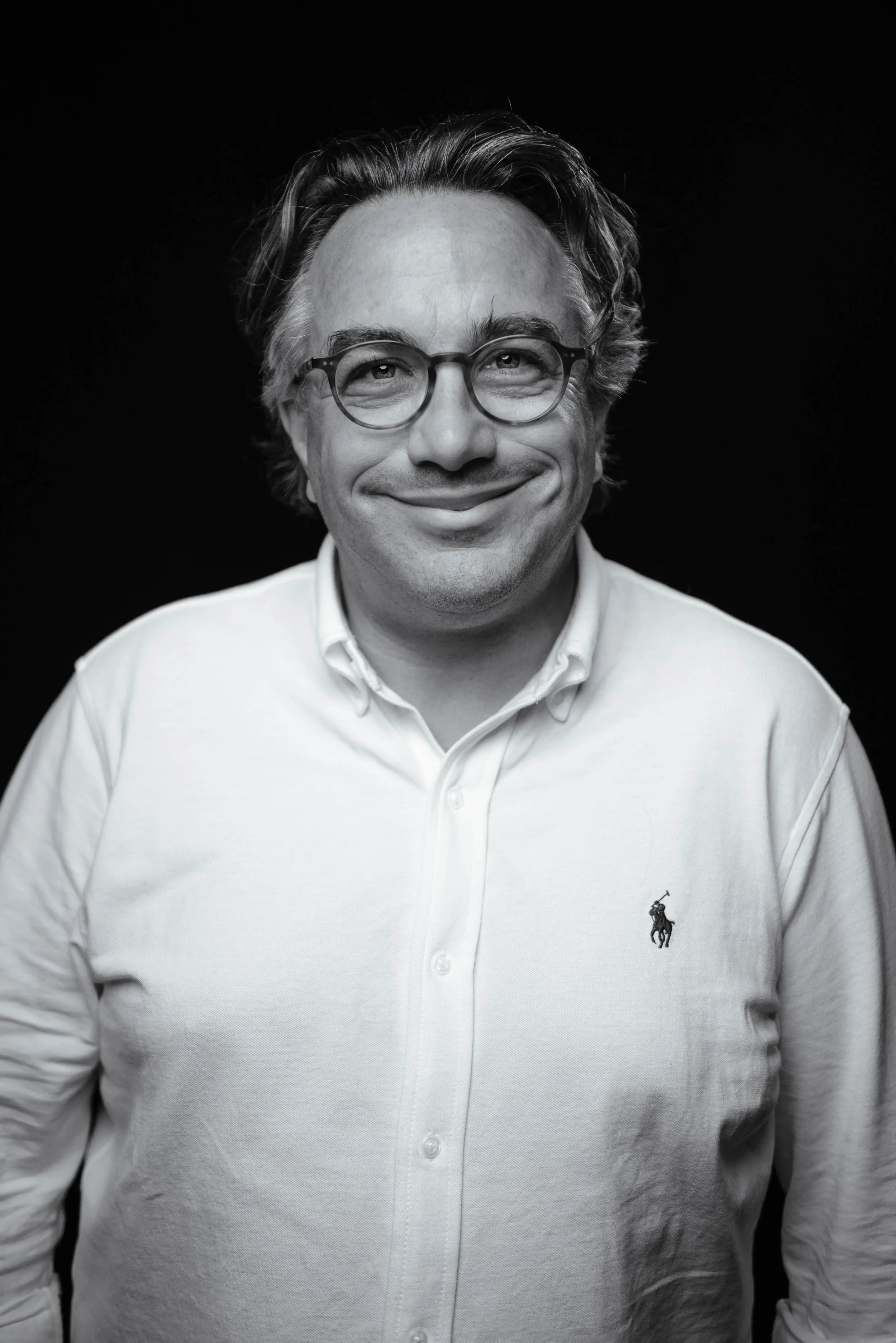 Porträt eines lächelnden Mannes mit Brille und weißem Hemd vor schwarzem Hintergrund.