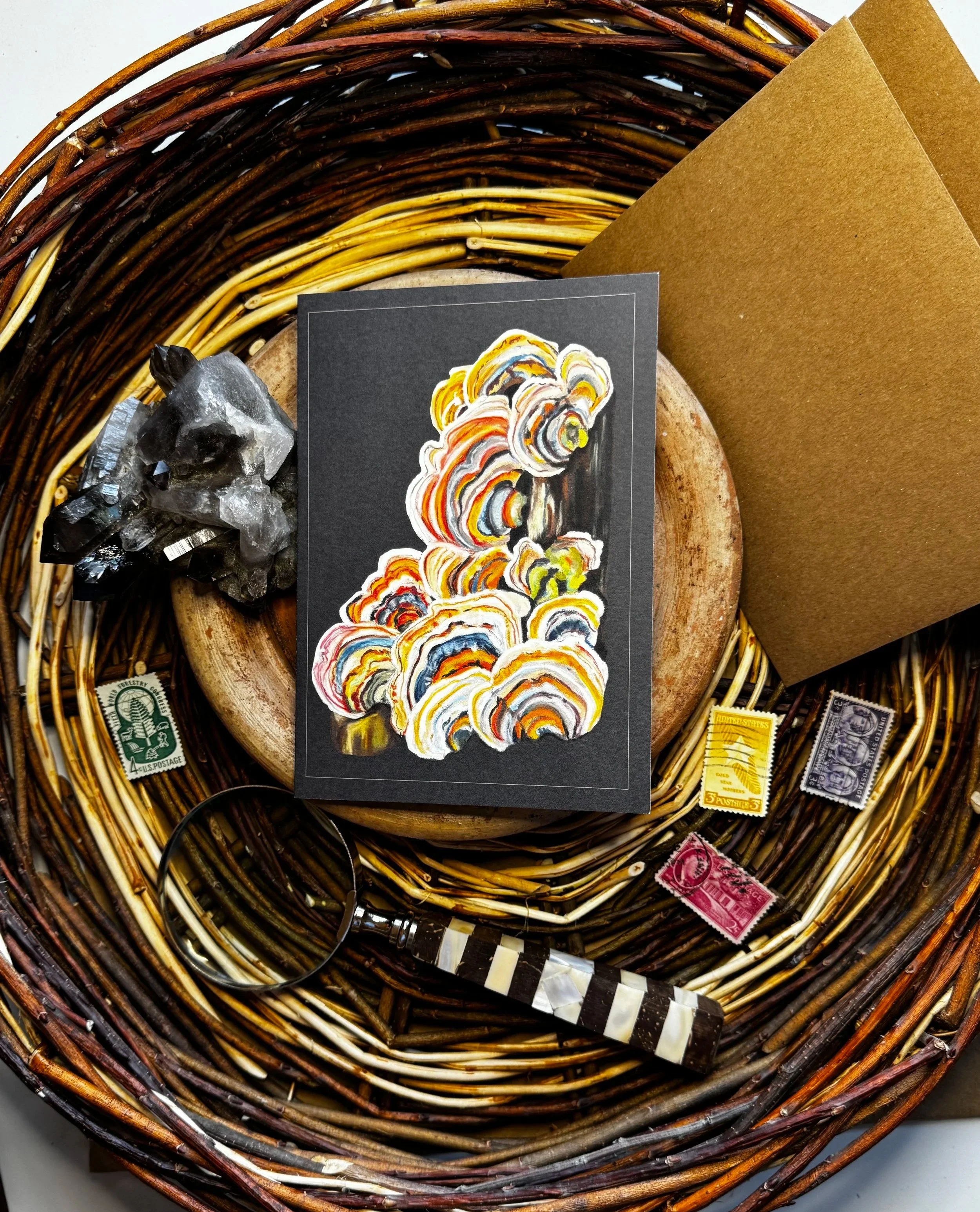A wicker basket containing various objects including a black and white striped object, several postage stamps, crystals, a piece of brown cardboard, a card with a colorful abstract design, and a glass