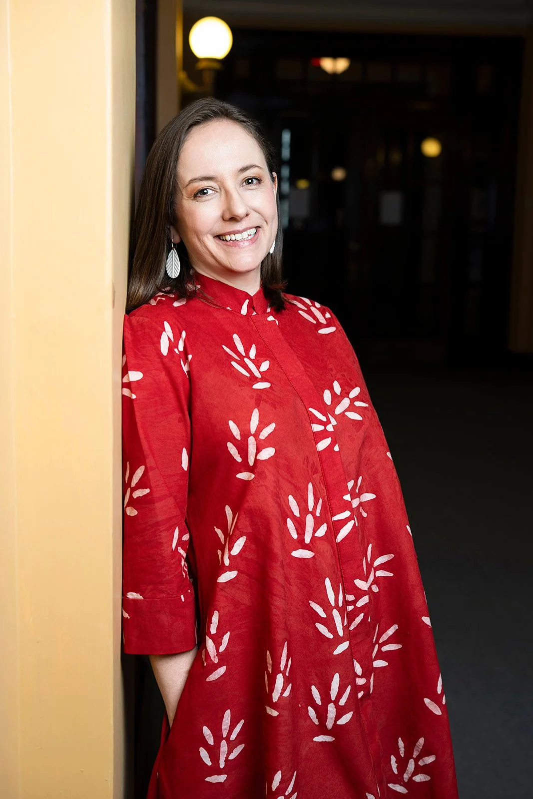 A woman with long brown hair wearing a red dress with white leaf patterns, smiling and leaning against a yellow wall in a dimly lit indoor setting.