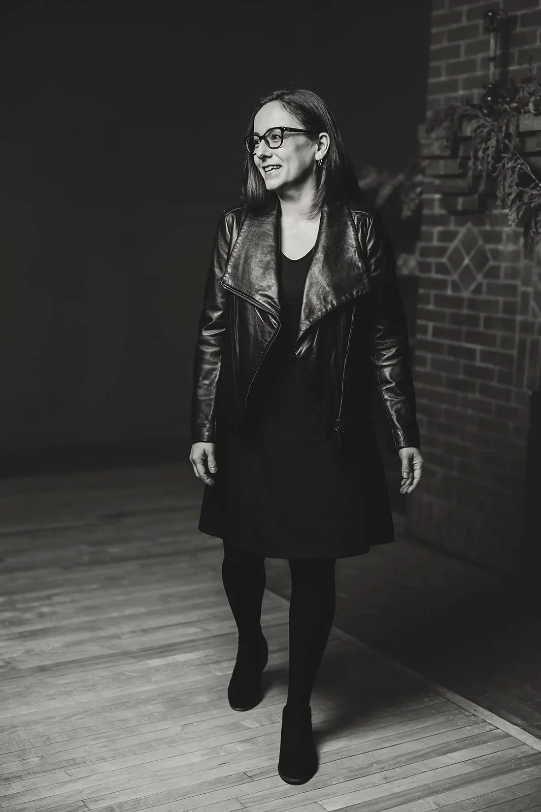 A woman wearing glasses, a leather jacket, and a dress walking in an indoor space with wooden flooring and a brick wall in the background.