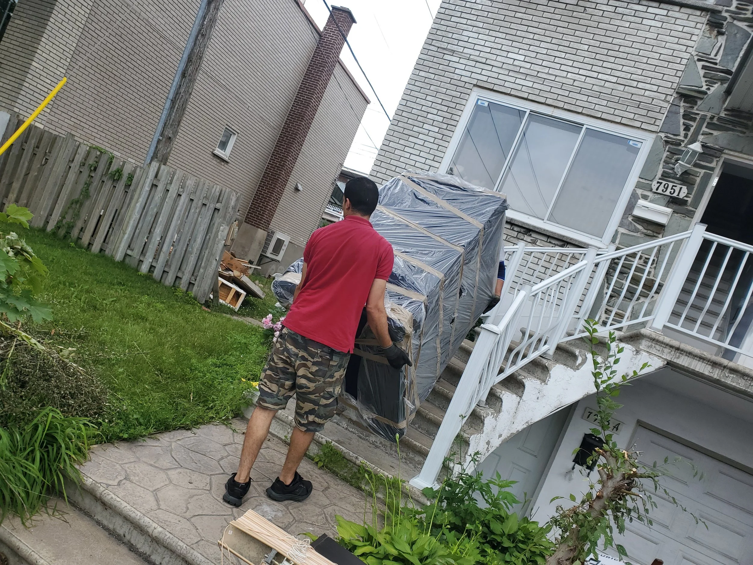 Un homme en t-shirt rouge et short camo transporte un objet emballé devant une maison. On voit un escalier extérieur, du gazon et une clôture en bois dans la scène.