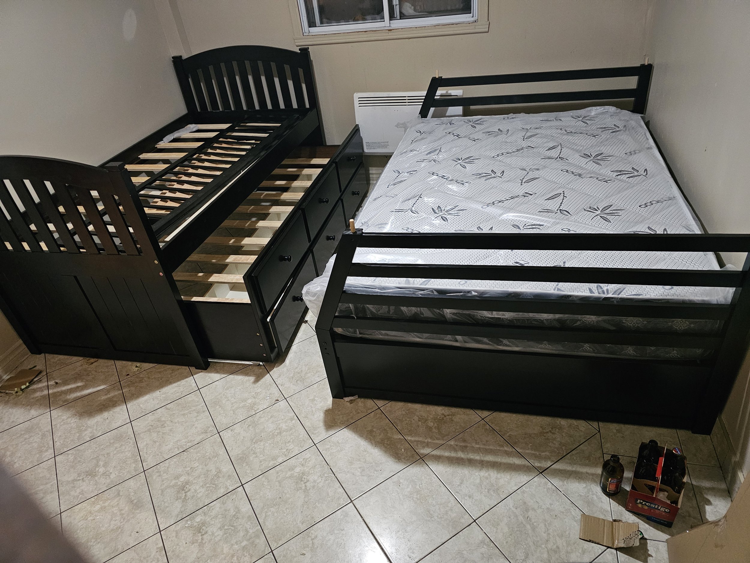 Deux lits noirs dans une pièce avec un sol en carrelage. L'un des lits a un espace de rangement avec des tiroirs en dessous. Le lit double est recouvert d'un matelas avec un motif de feuilles. Il y a aussi un pack de bouteilles vides sur le sol.
