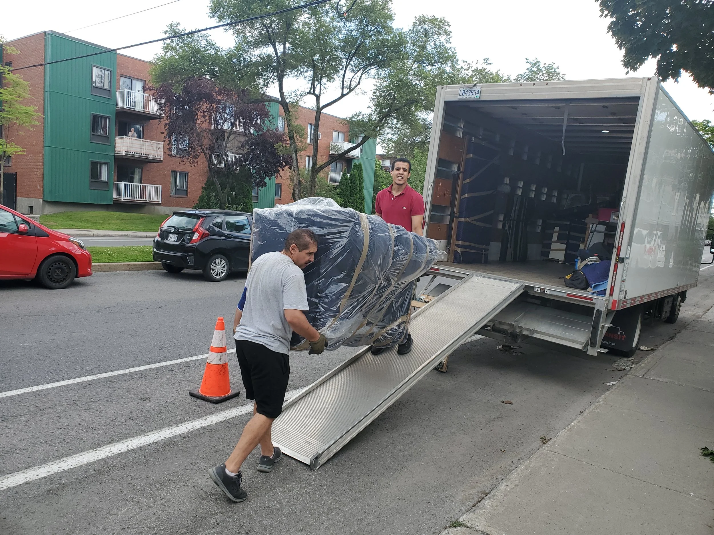 Deux hommes déchargent un grand meuble enroulé dans du plastique depuis un camion de déménagement garé sur une rue résidentielle. Un cône orange est placé à côté du camion. On voit des immeubles résidentiels et des voitures en arrière-plan.