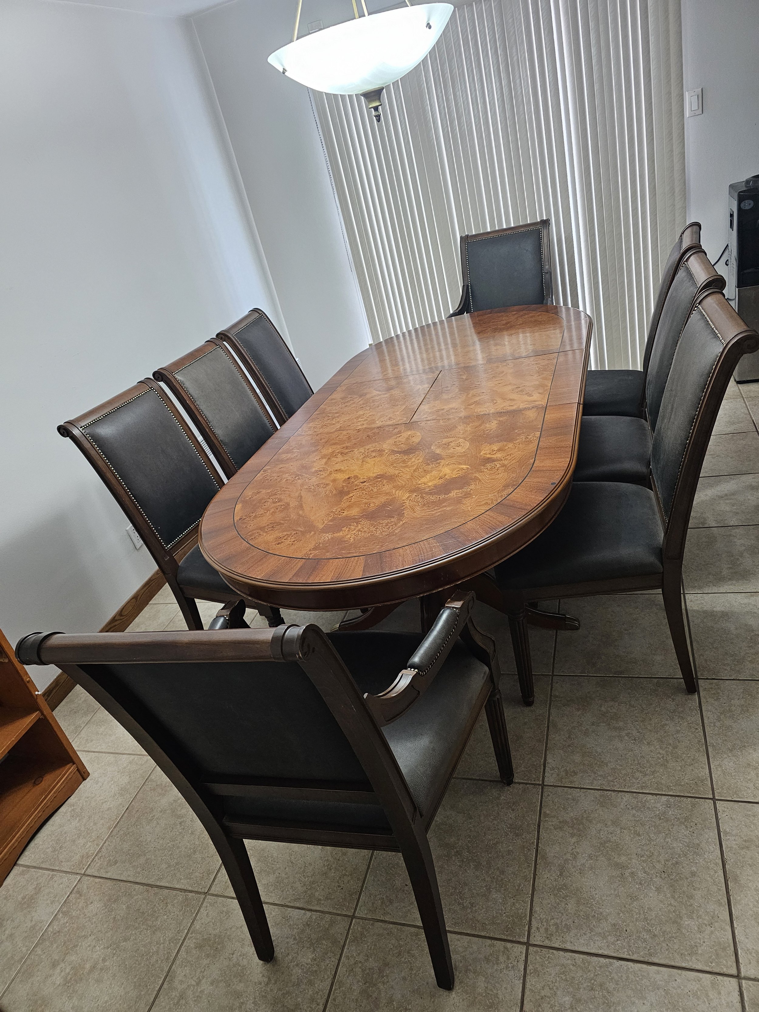 Salle à manger avec une table en bois ovale et huit chaises, un luminaire suspendu et des stores verticaux en arrière-plan.