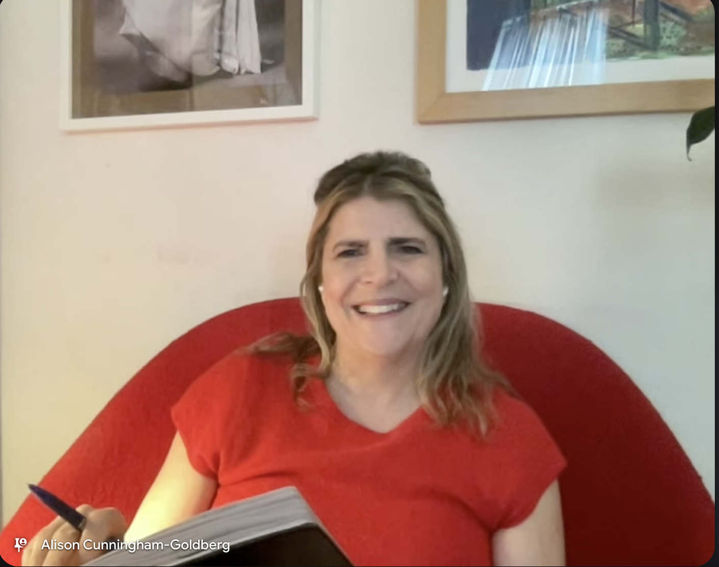 A woman with shoulder-length blonde hair, smiling, wearing a red top, sitting on a red chair in a room with framed pictures on the wall behind her. She appears to be holding a pen in her right hand and a notebook in her lap.