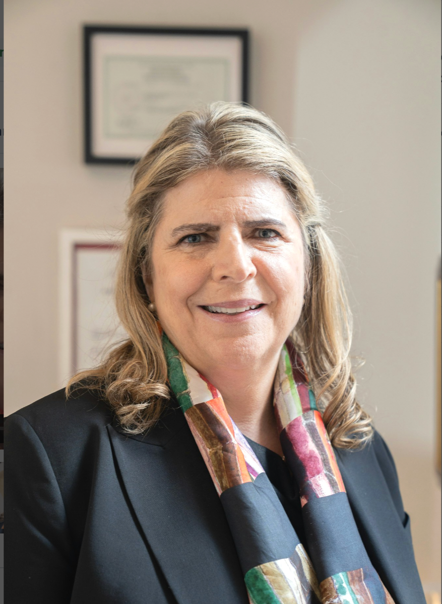 A portrait of a middle-aged woman with blonde hair, smiling, wearing a black blazer and a colorful scarf, standing in an office or professional setting with framed documents on the wall behind her.