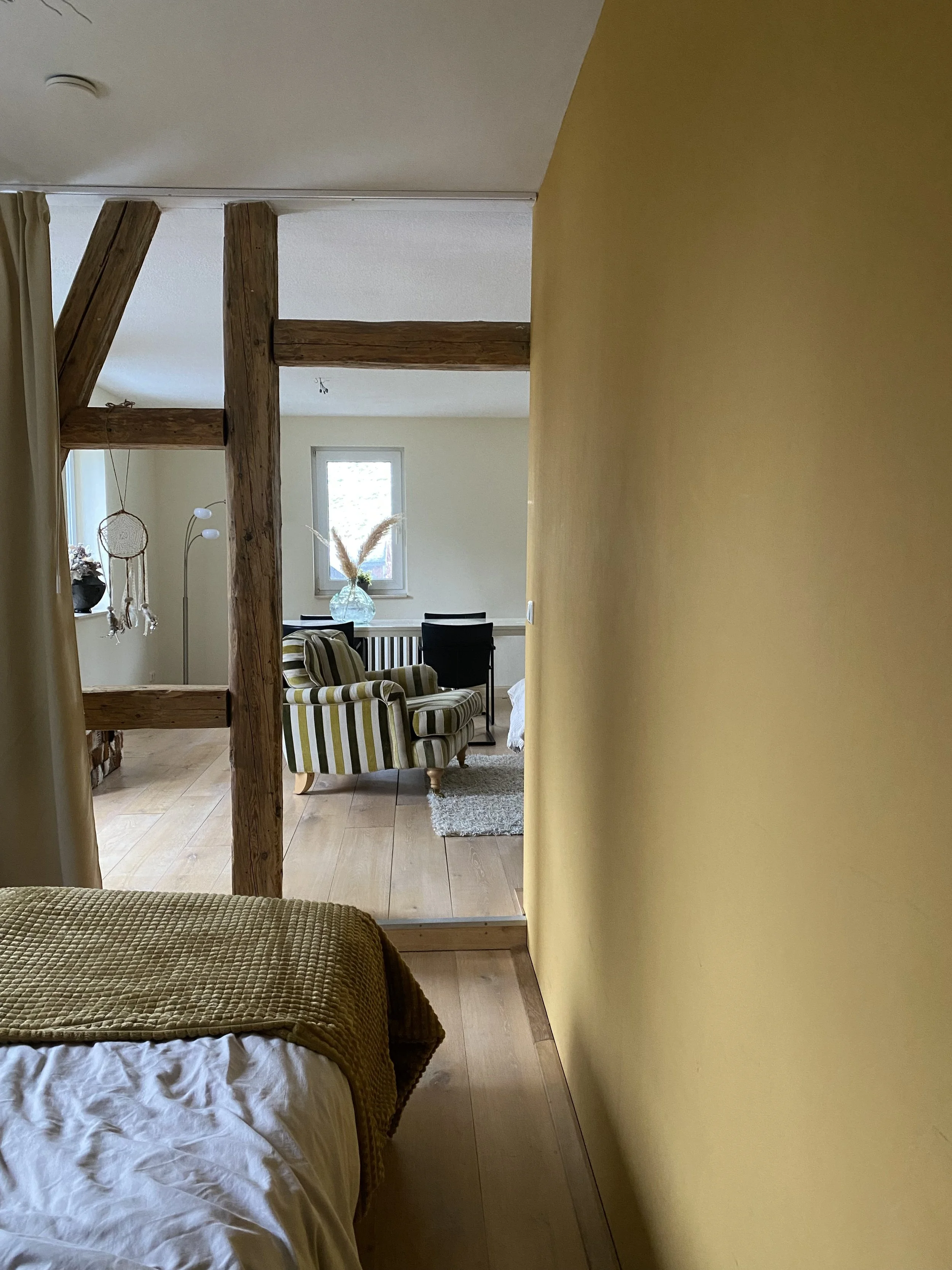 Ein Blick in ein Zimmer mit einem gelben Wandabschnitt, Holzbalken, einem Bett mit gelber Decke, einem Sessel mit grünen, weißen und braunen Streifen, einem kleinen Teppich und einem Schreibtisch im Hintergrund.