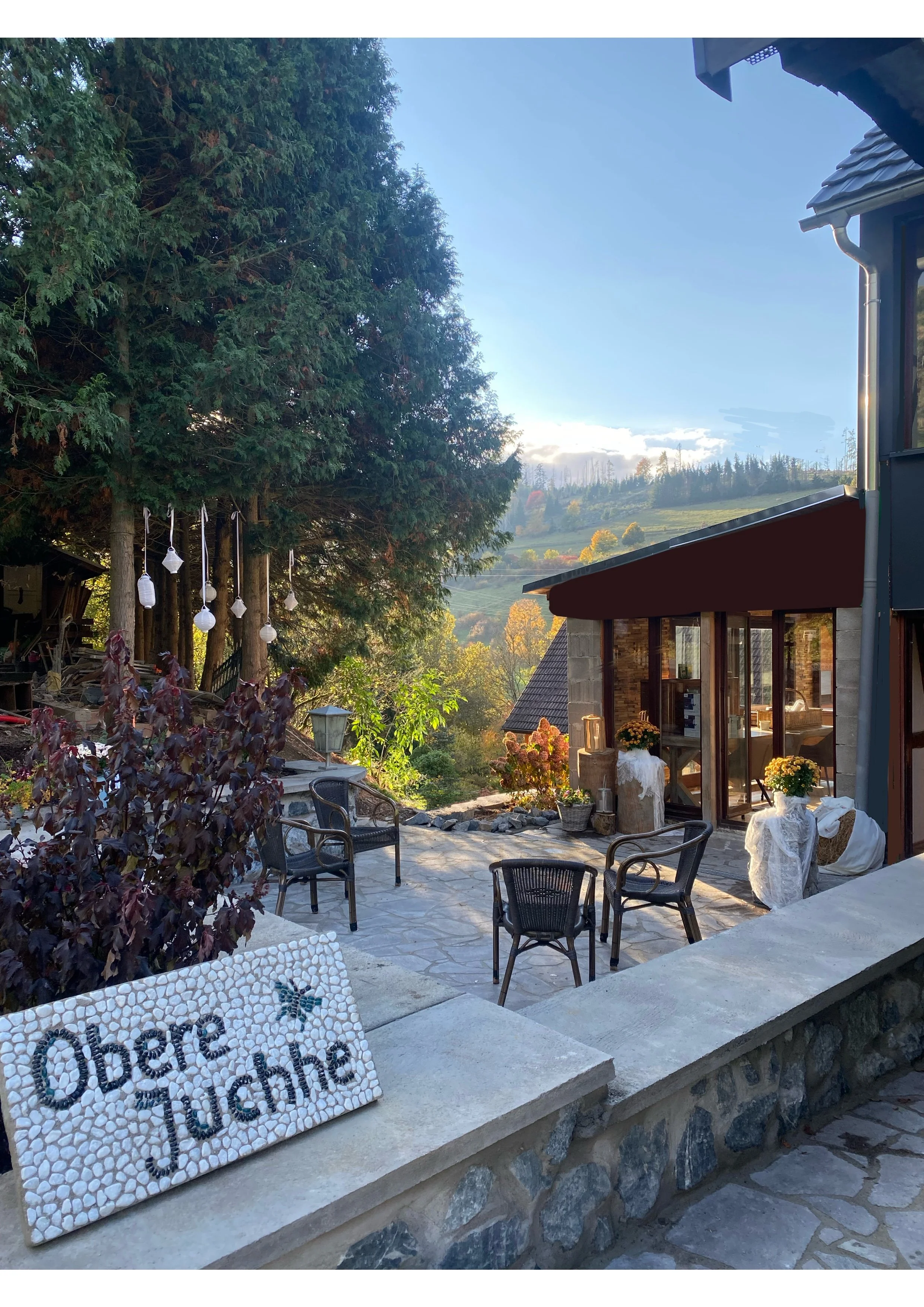 Gemütlicher Außenbereich eines Hauses mit mehreren Stühlen, Töpfen mit Blumen und einem Schild mit der Aufschrift 'Oberre Juchhe' inmitten von Bäumen und einer bergigen Landschaft im Hintergrund.