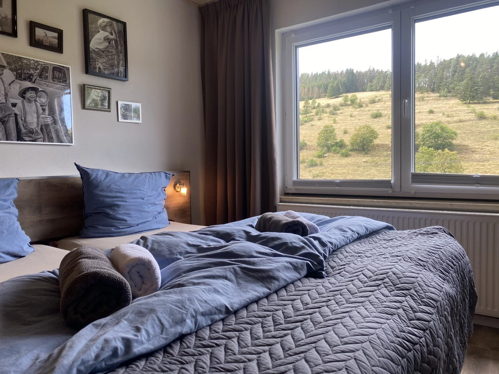 Schlafzimmer mit Bett, Blaue Kissen, gerollte Handtücher, Wandbilder und großes Fenster mit Blick auf eine grüne hügelige Landschaft