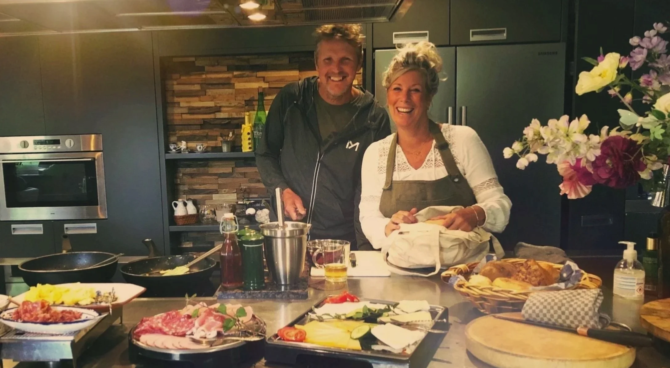 Zwei lachende Personen, ein Mann und eine Frau, stehen in einer modernen Küche neben einem Herd und einer Arbeitsplatte mit verschiedenen Zutaten und Küchenutensilien. Im Hintergrund sind Regale mit Geschirr und eine Wand mit flacher Steinverkleidung. Neben ihnen sind Blumen in einer Vase und verschiedene Lebensmittel auf der Theke zu sehen.