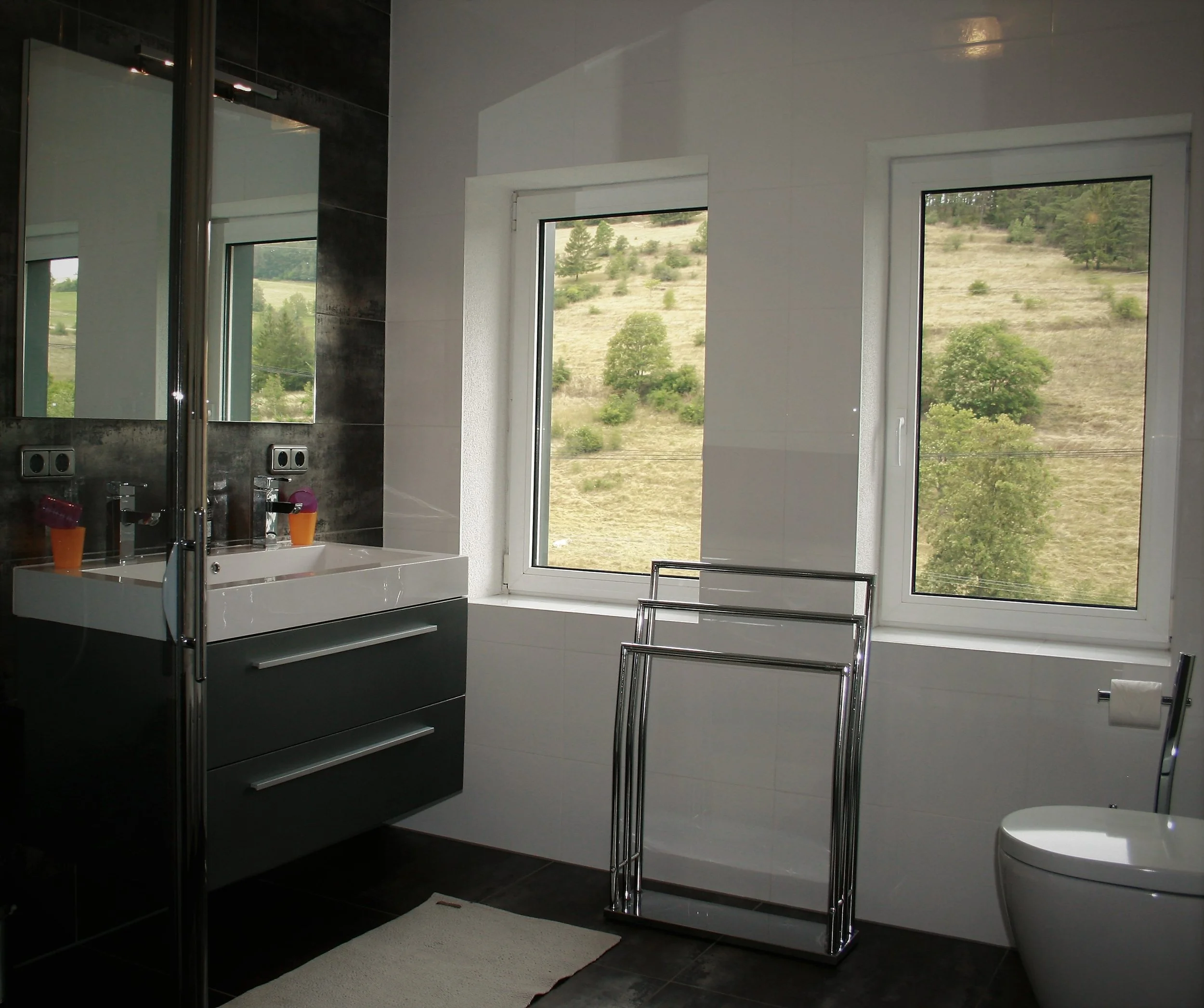 Ein modernes Badezimmer mit dunklem Fliesenspiegel, weißem Waschbecken, zwei großen Fenstern mit Blick auf eine grüne Berglandschaft, einer weißen Toilette, einem Handtuchständer und einer weißen Badevorlage auf dem Boden.