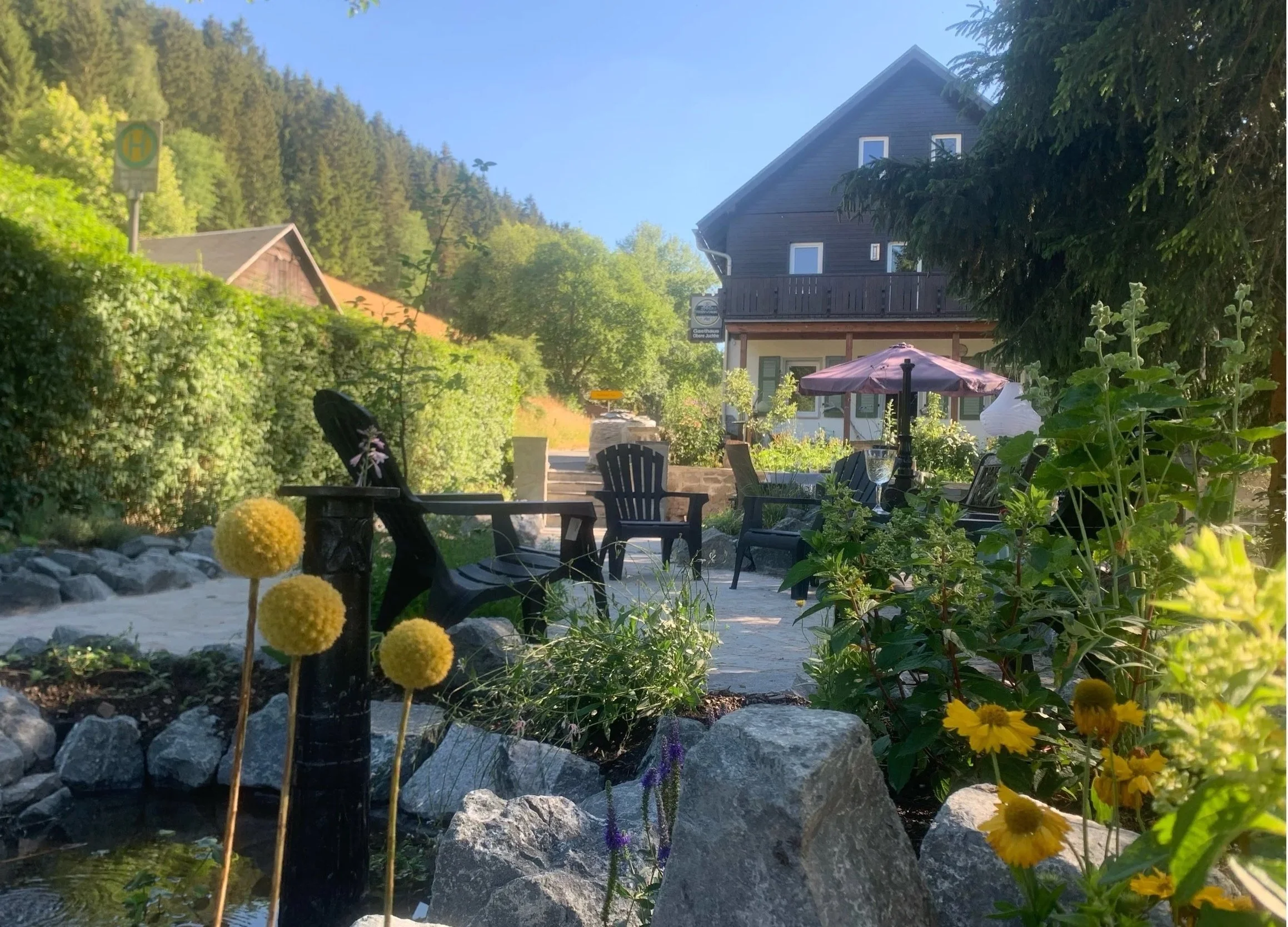 Gemütlicher Garten mit Gartenmöbeln, Pflanzen, Sonnenstrahlen, ein Haus im Hintergrund, und Berge im Himmel.