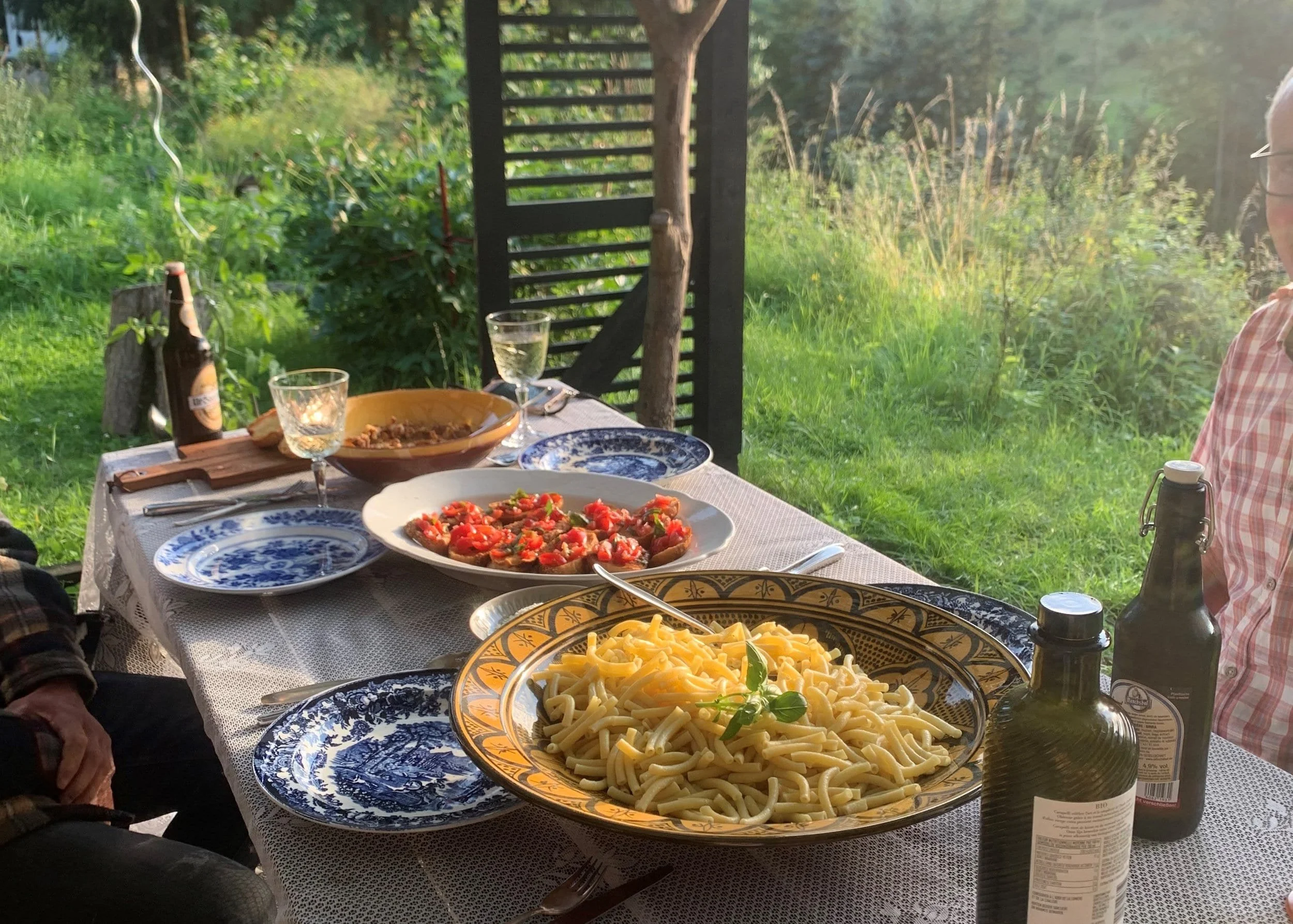 Ein gedeckter Tisch mit Pasta, Tomaten und weiteren Gerichten im Garten bei Sonnenuntergang, umgeben von grüner Natur und Bäumen.