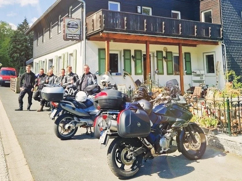 Eine Gruppe von Motorradfahrern steht vor einem Gebäude mit Balkon und Garten im Hintergrund, mehrere Motorräder parken davor.