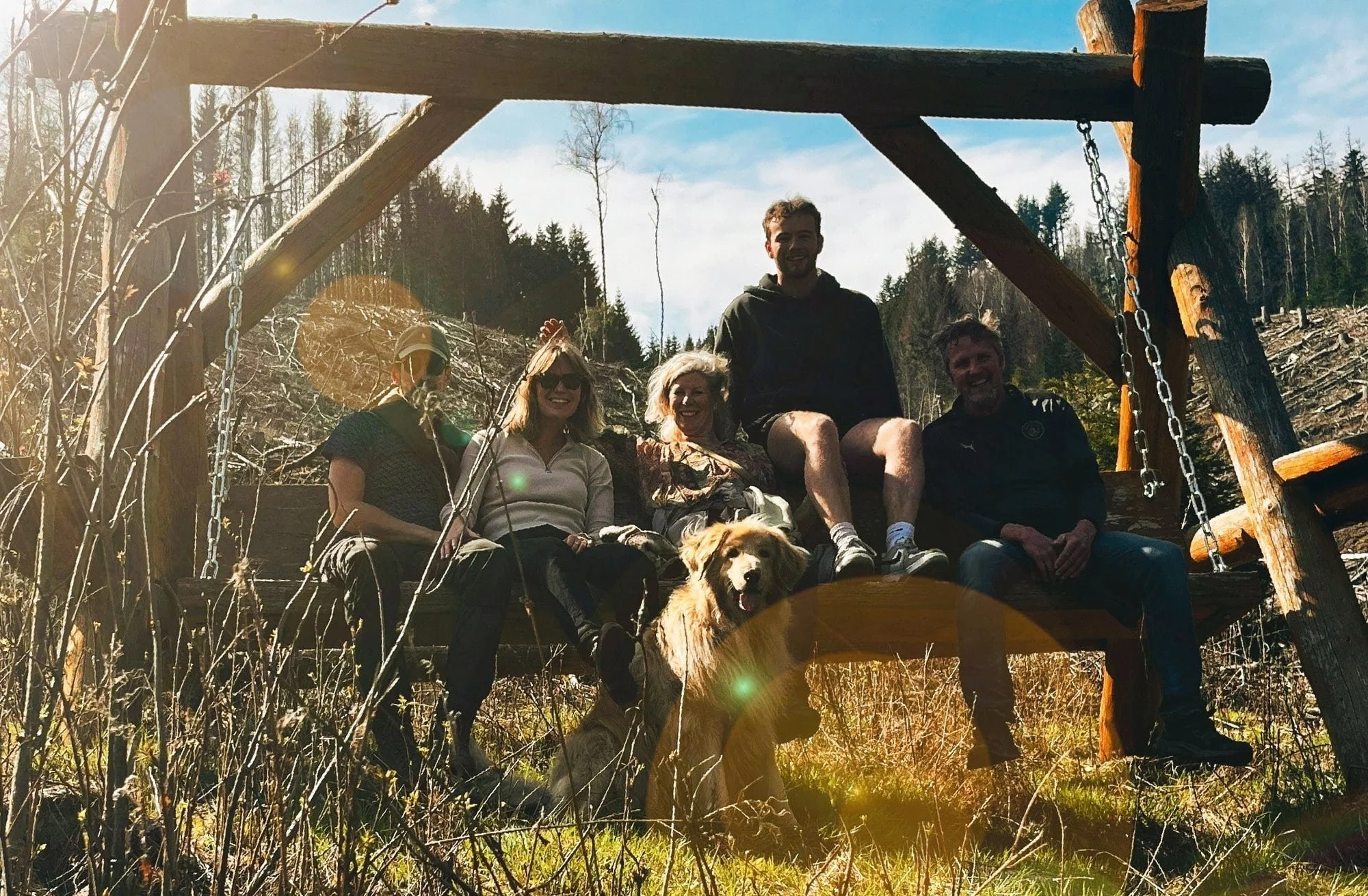 Eine Gruppe von sechs Menschen sitzt auf einer Baum-Schaukel im Freien, zusammen mit einem Hund, umgeben von Bäumen und Bergen bei sonnigem Wetter.