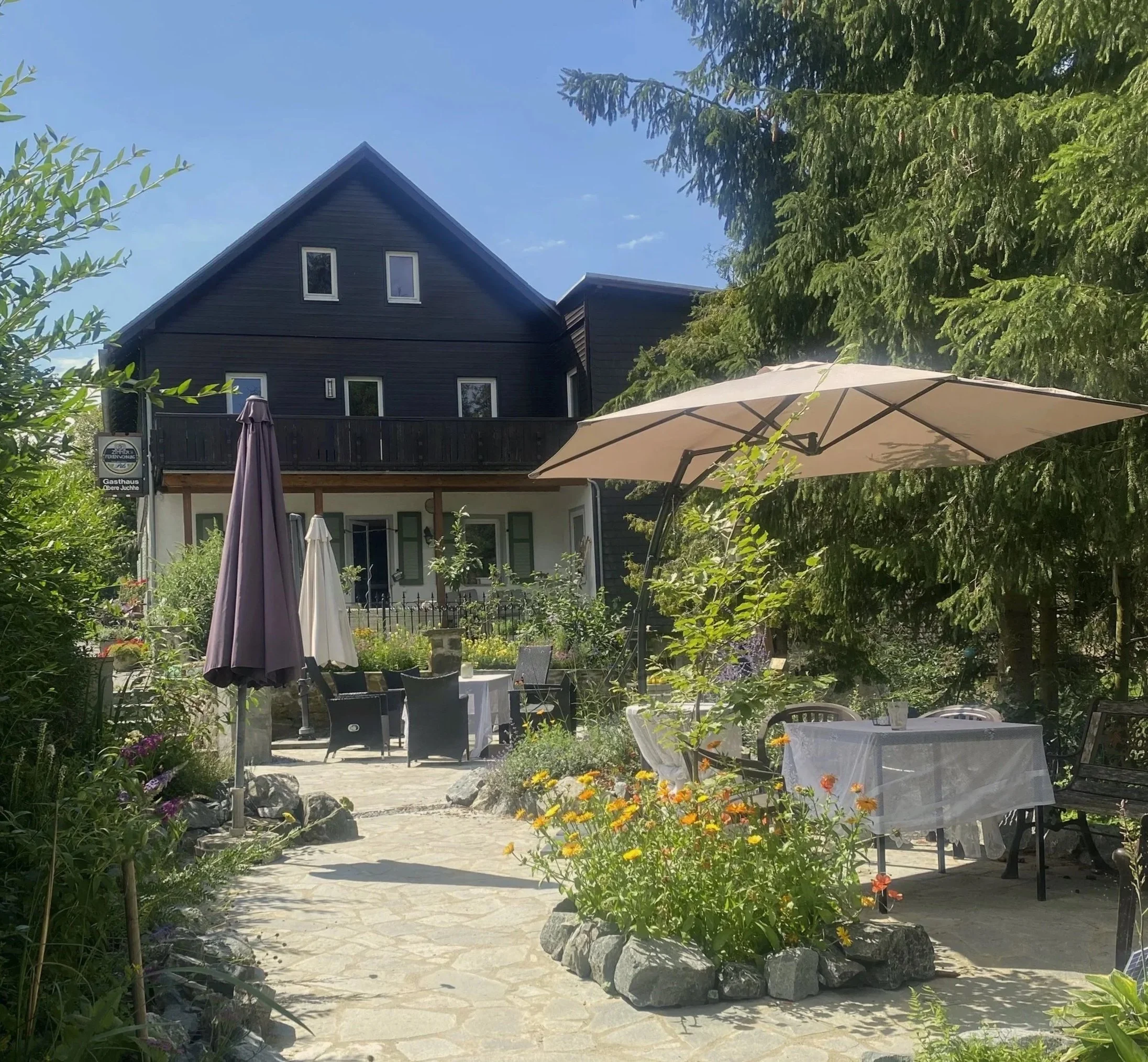 Ein Außenbereich mit Tischen, Stühlen und Sonnenschirmen vor einem schwarzen Haus mit Balkon, umgeben von Gartenpflanzen und Blumen. Es ist sonnig und der Himmel ist blau.