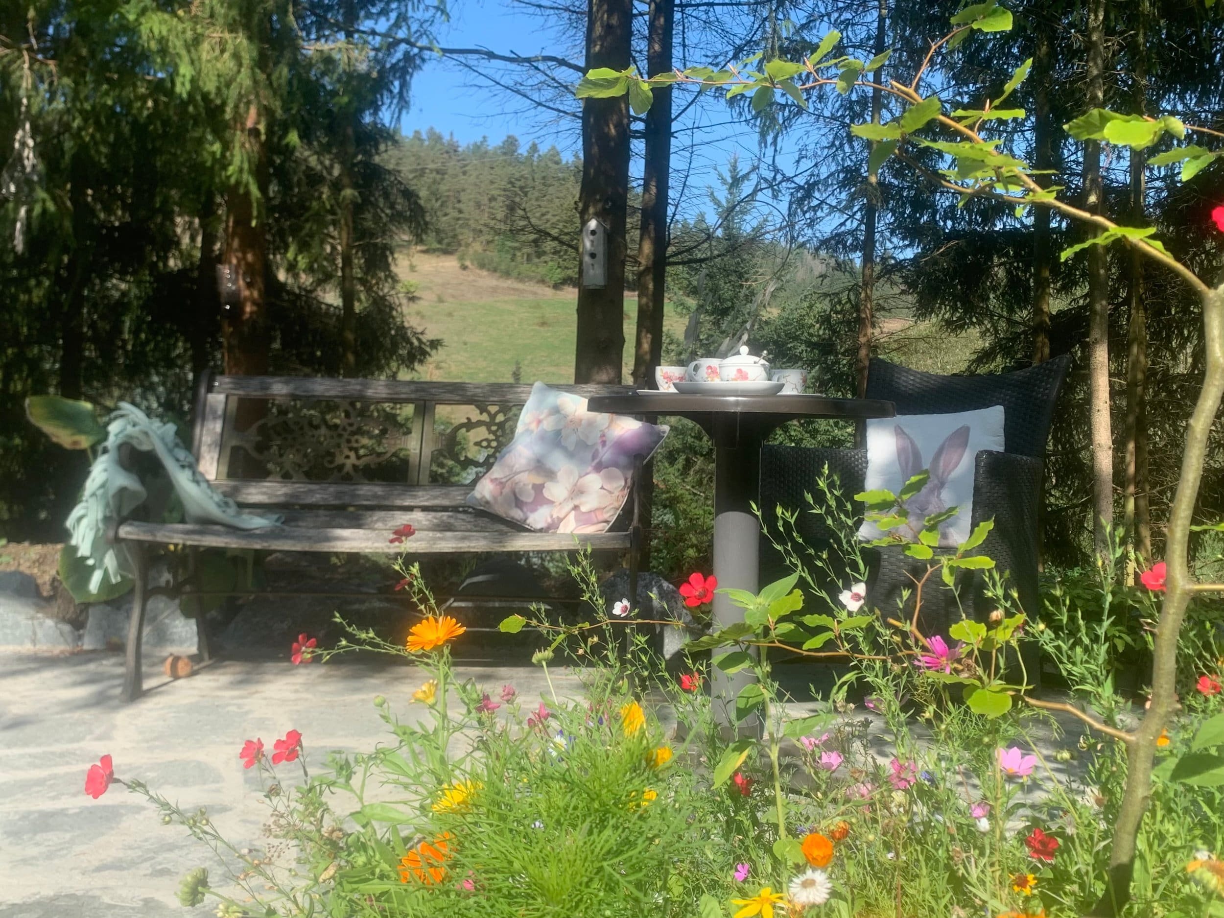 Ein Garten mit einer Holzgartenbank, einem Tisch und zwei Stühlen, umgeben von bunten Blumen und Bäumen, bei sonnigem Wetter.