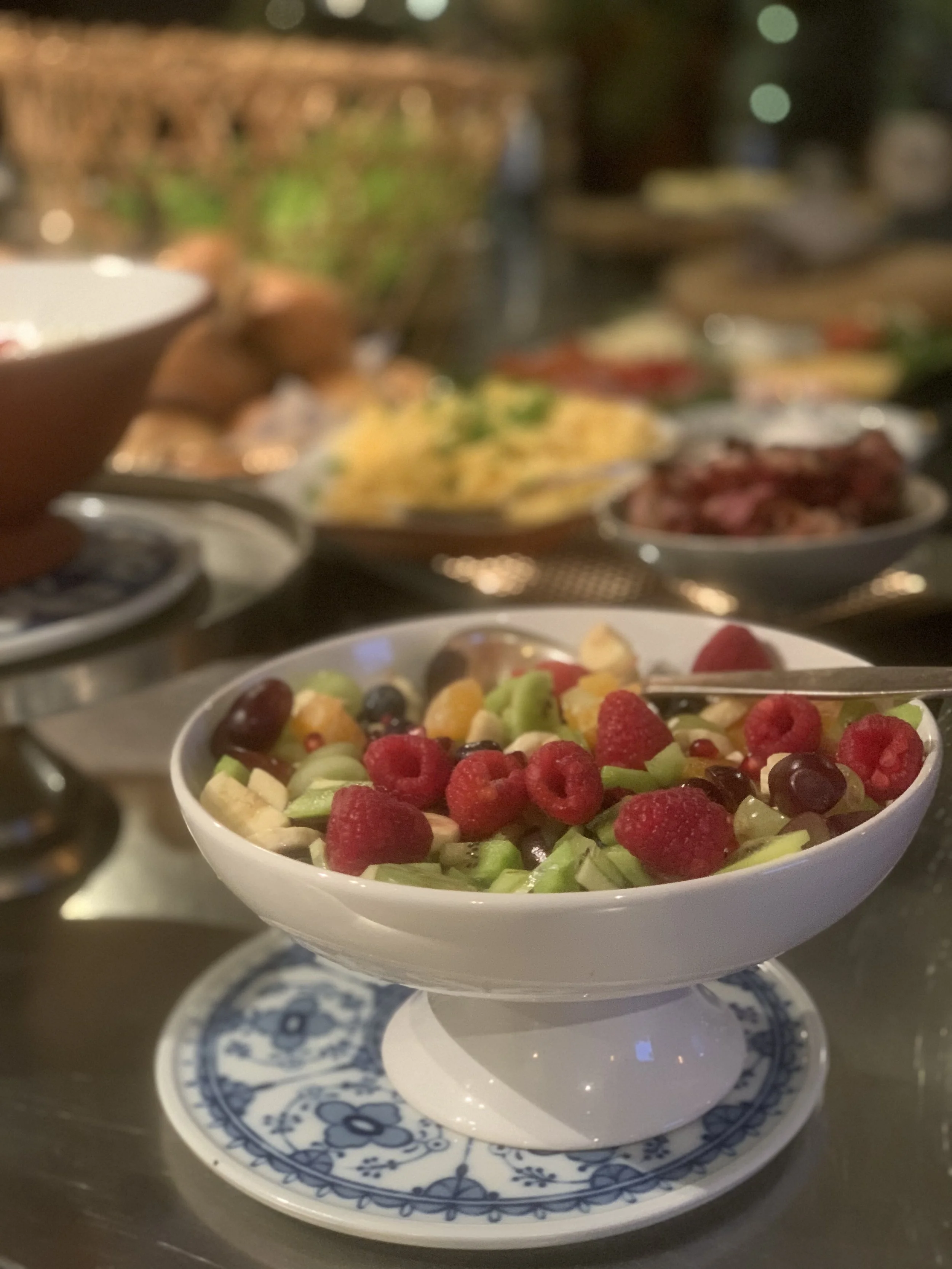 Schüssel mit gemischtem Obstsalat, umgeben von verschiedenen Tellern mit Speisen auf einem gedeckten Tisch.
