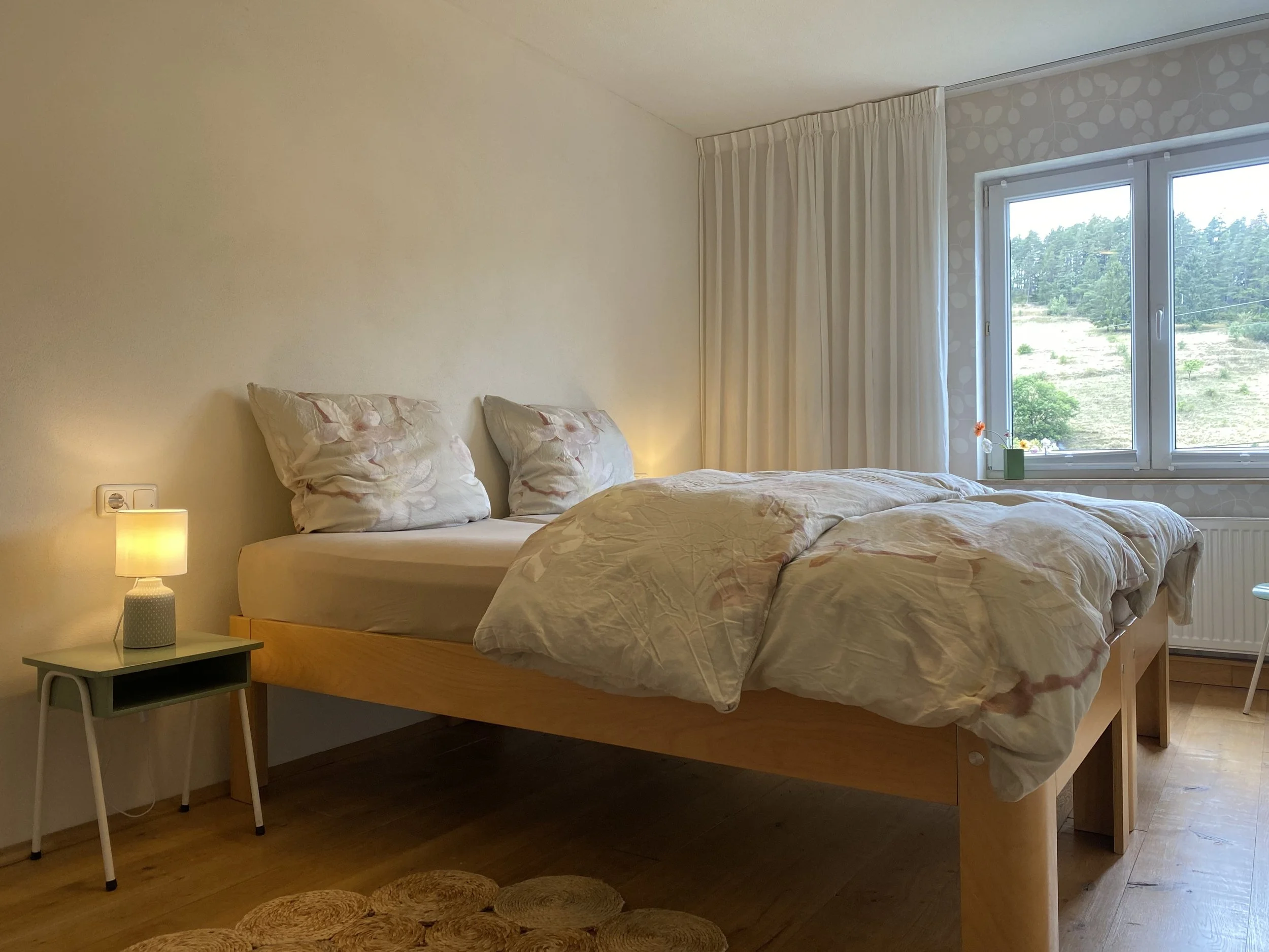 Schlafzimmer mit einem Bett, einem Nachttisch mit Lampe, einem Fenster mit Blick auf eine grüne Landschaft, weißen Vorhängen und Holzfußboden.