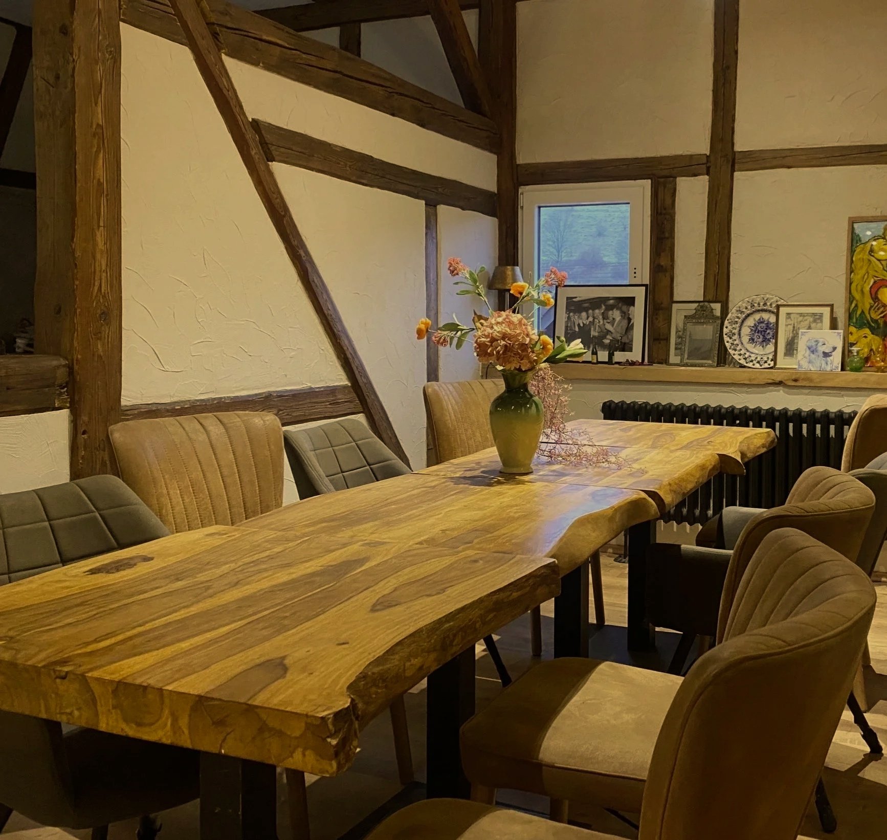 Rustikaler Esszimmerbereich mit einem großen hölzernen Tisch, umgeben von gepolsterten Stühlen und einer Vase mit Blumen in der Mitte, im Hintergrund Bilder und Dekorationen auf einem Fensterbank, in einem Raum mit Holzbalken und -wänden.