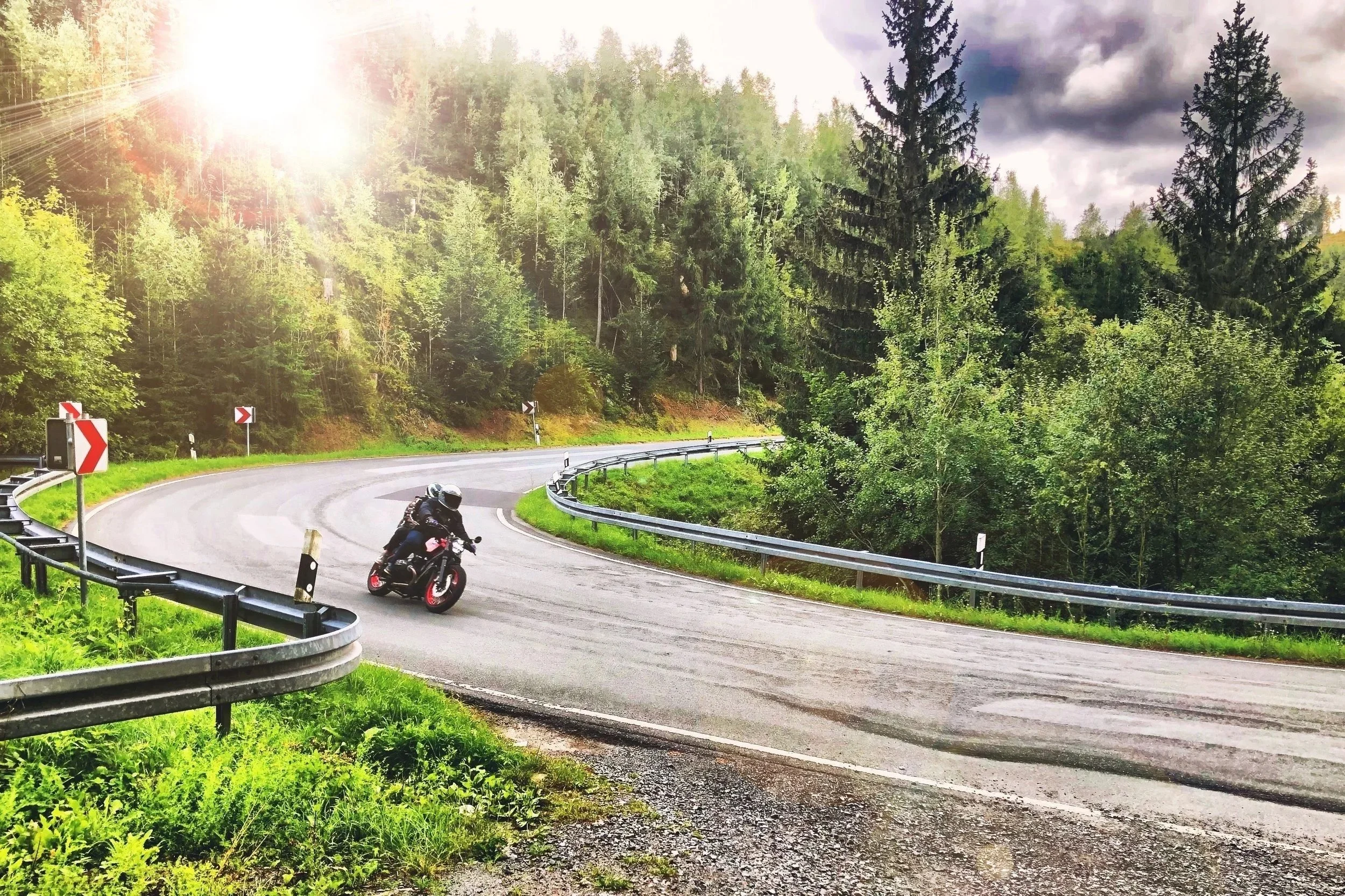 Motorradfahrer auf kurviger Bergstraße inmitten von grünen Bäumen, mit Sonnenlicht und dunklen Wolken am Himmel.