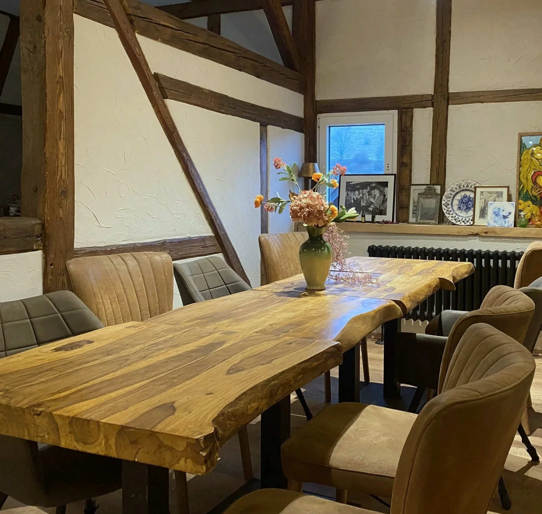 Ein Holztisch in einem gemütlichen Raum mit antiken Stühlen, einer Vase mit Blumen in der Mitte, einem Fenster im Hintergrund und verschiedenen Bildern und Dekorationen auf einem Fensterbrett.