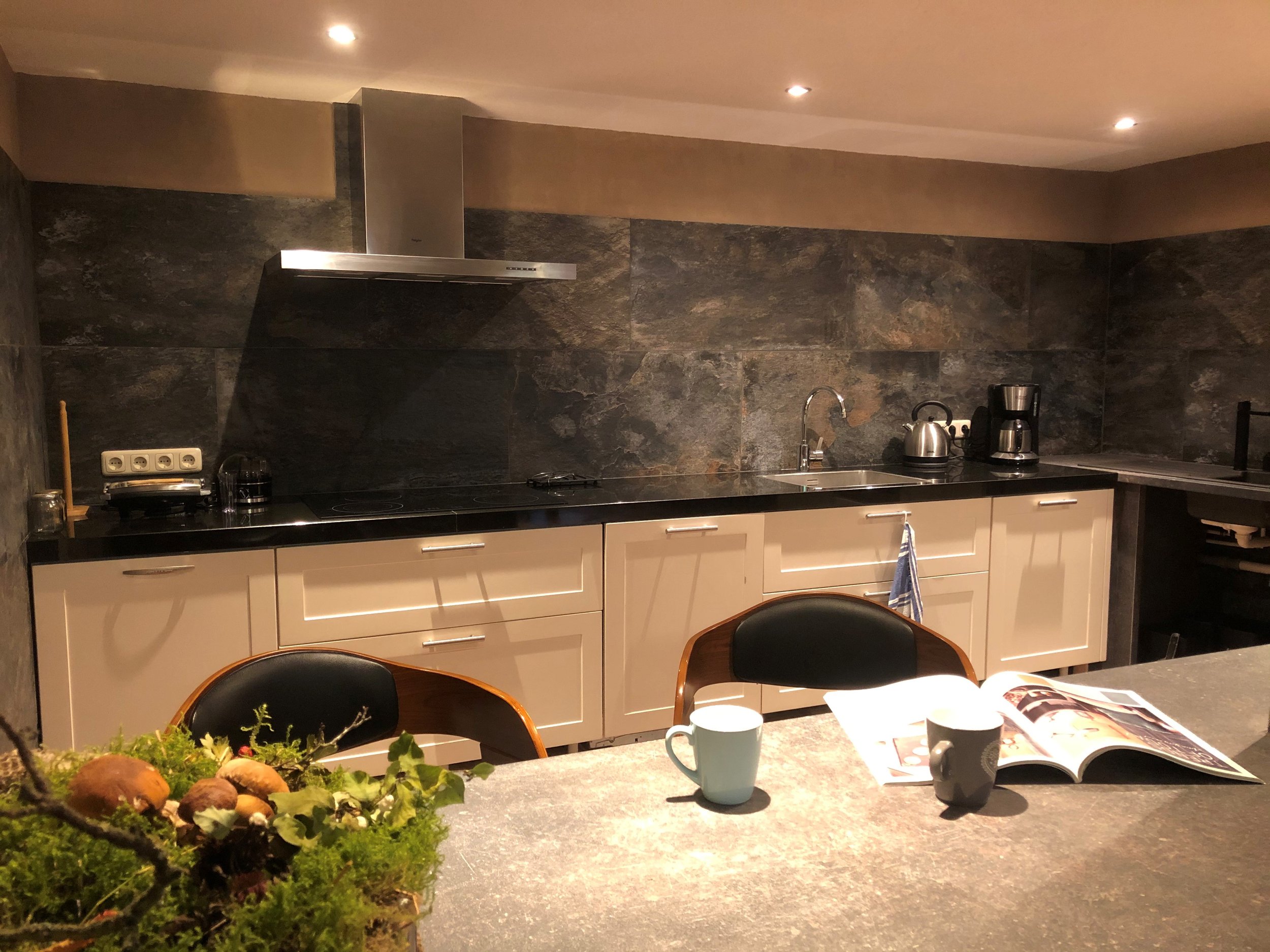 Küche mit schwarzen Arbeitsflächen, weißen Schränken und dunklen Steinwänden, zwei Tassen, eine Zeitschrift und eine Topfpflanze auf der Theke.
