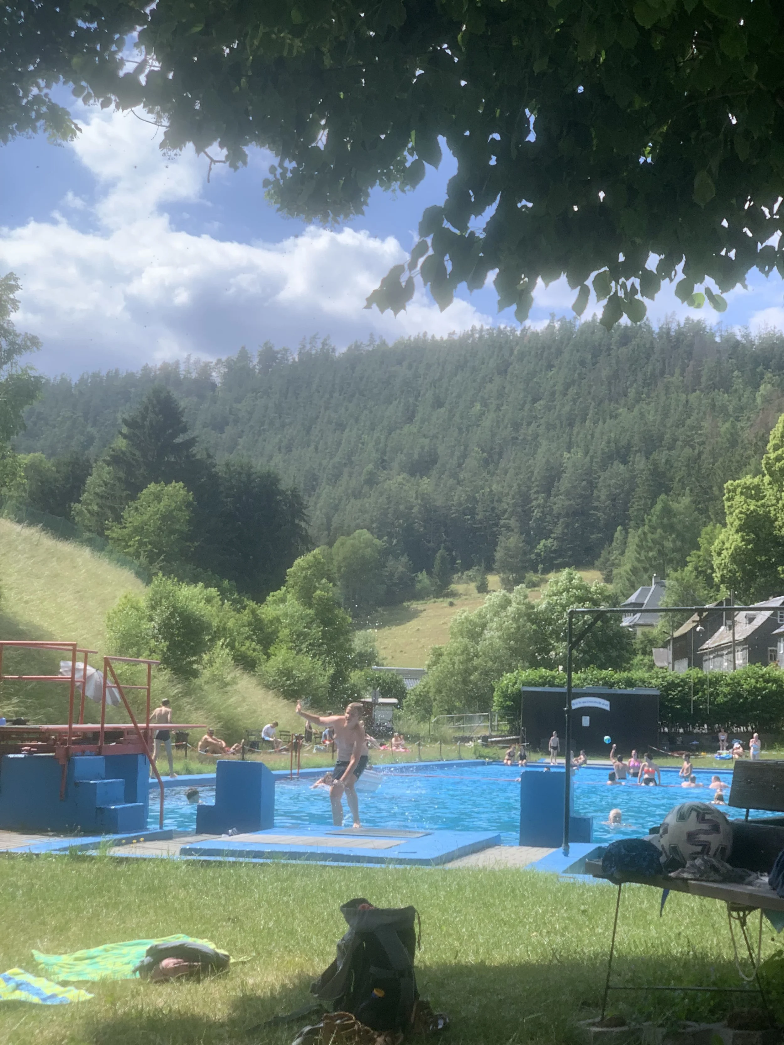 Ein Freibad mit mehreren Personen, die im Wasser schwimmen und spielen, umgeben von grünen Bäumen und Hügeln, bei sonnigem Wetter.