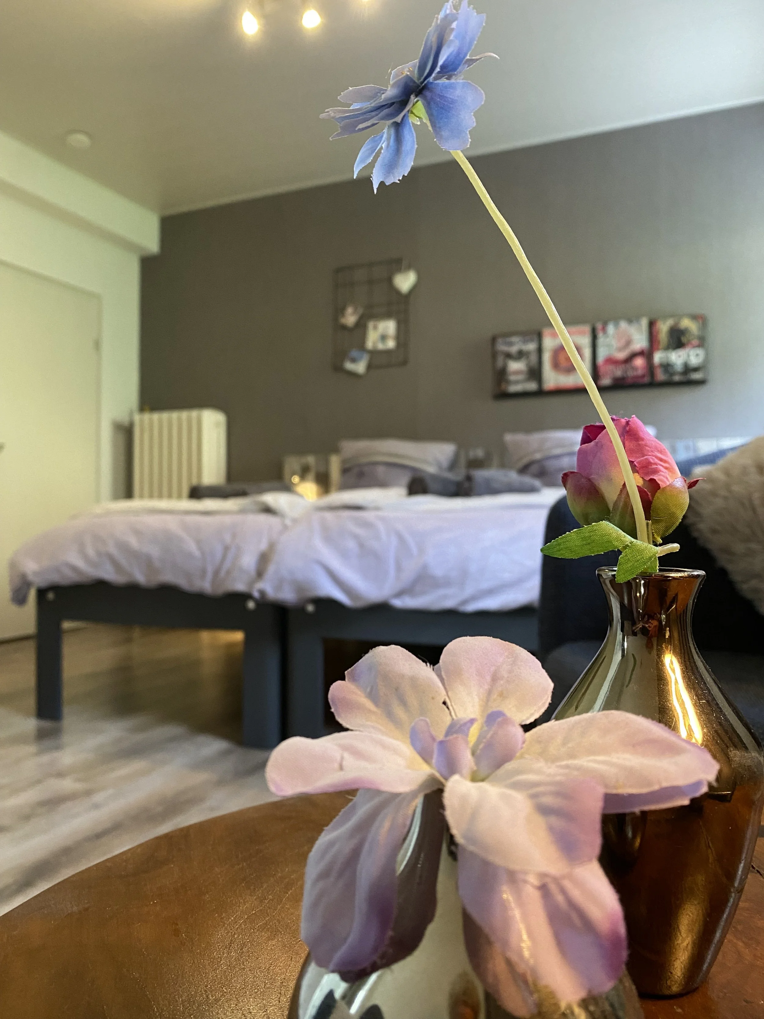 Blumen in einer kleinen Vase auf einem Tisch, im Hintergrund ein Schlafzimmer mit Betten und Dekorationen an der Wand.