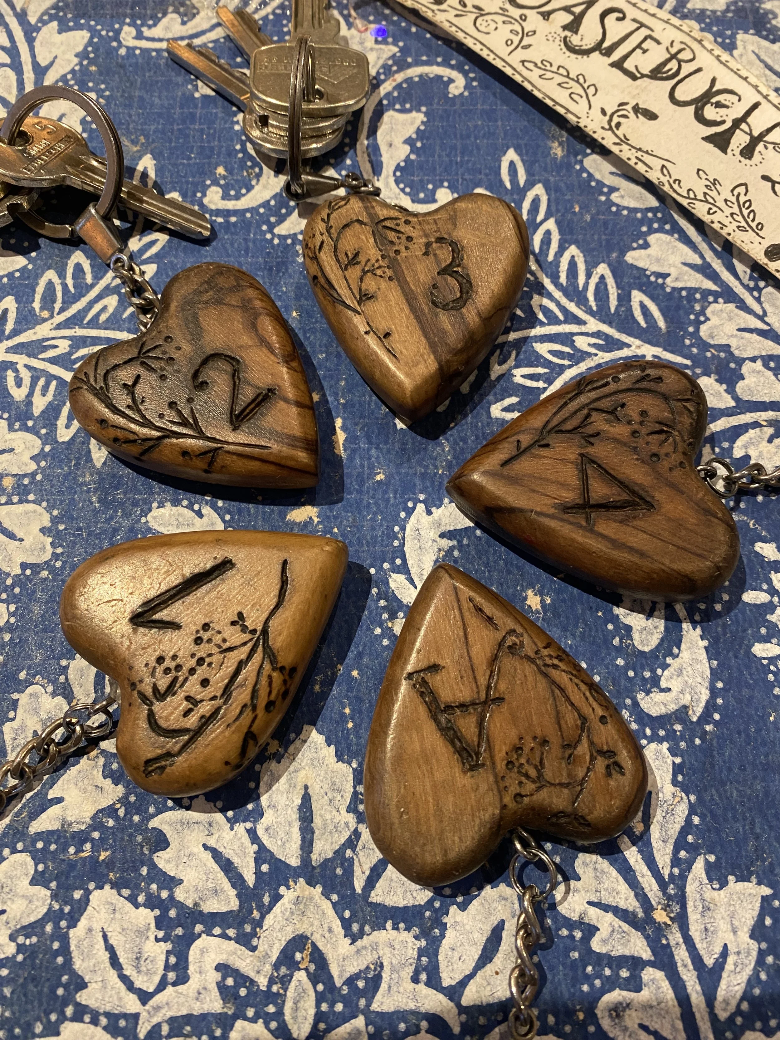 Sechs Holzherz-Schlüsselanhänger mit gravierten Zahlen, auf blau-weißem Musterhintergrund, neben Schlüssel und einer Zeitung.