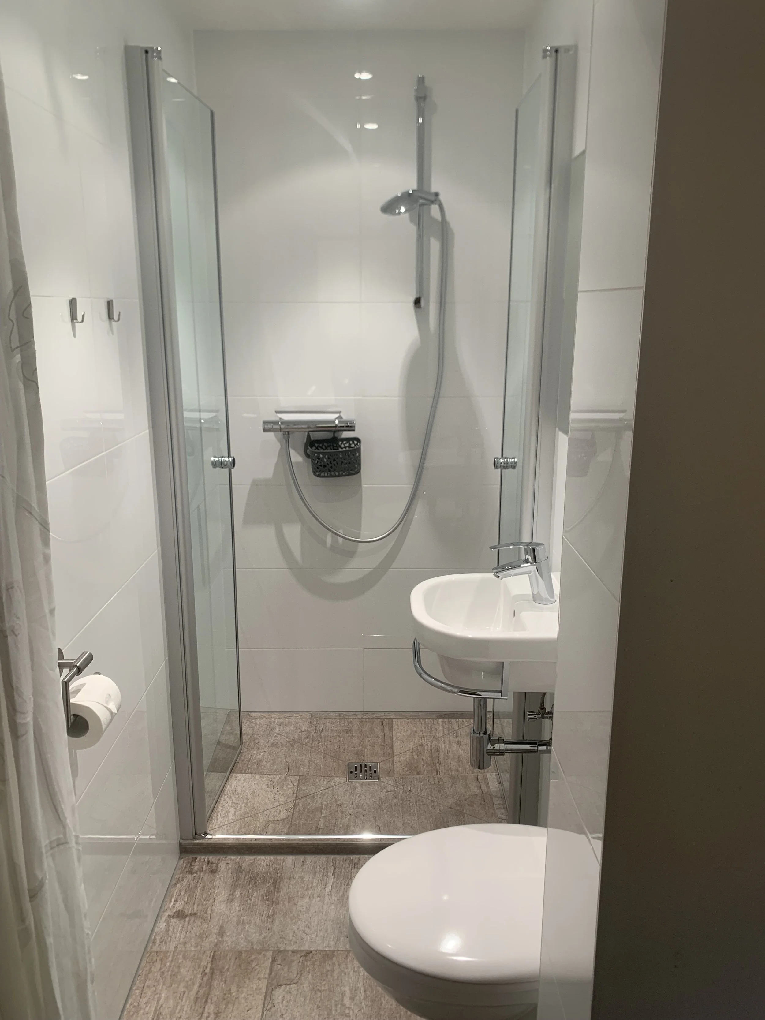 Kleines Badezimmer mit Dusche, Waschbecken und Toilette, weiß gefliest, mit grauem Bodenbelag.